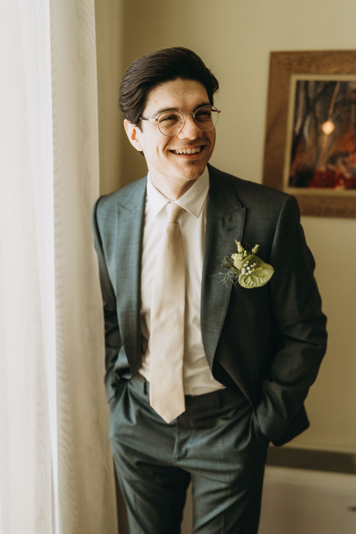 Smiling groom in dark suit with boutonniere and cream tie stands by window on wedding day.