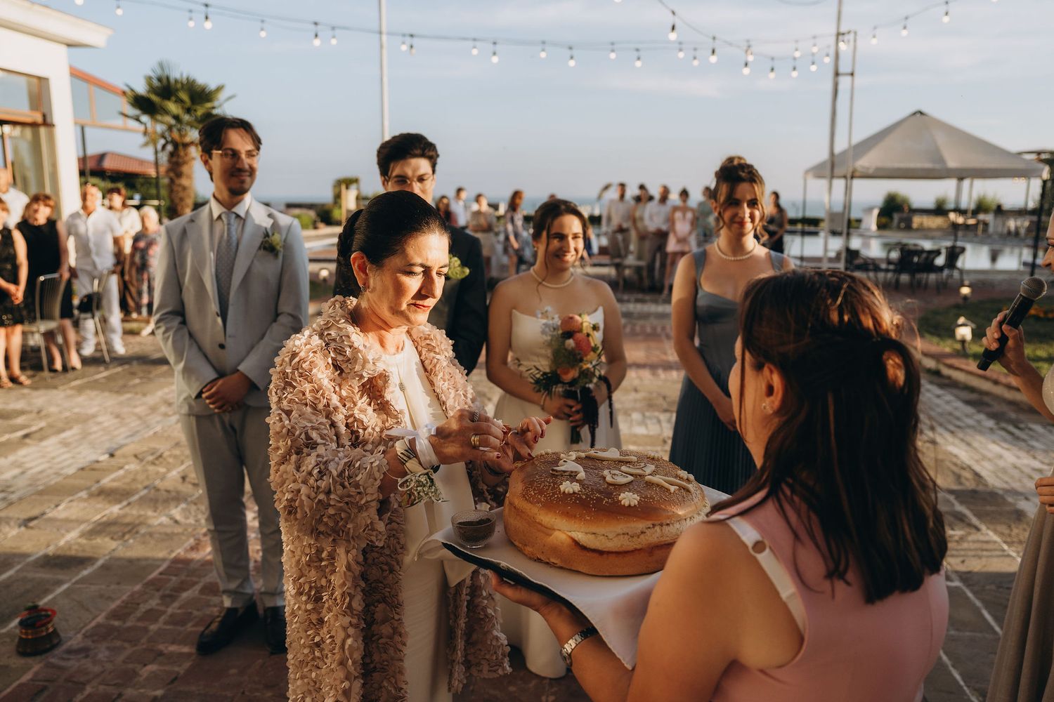 Outdoor wedding ceremony with guests watching as a woman in a floral shawl participates in a ring exchange at sunset.