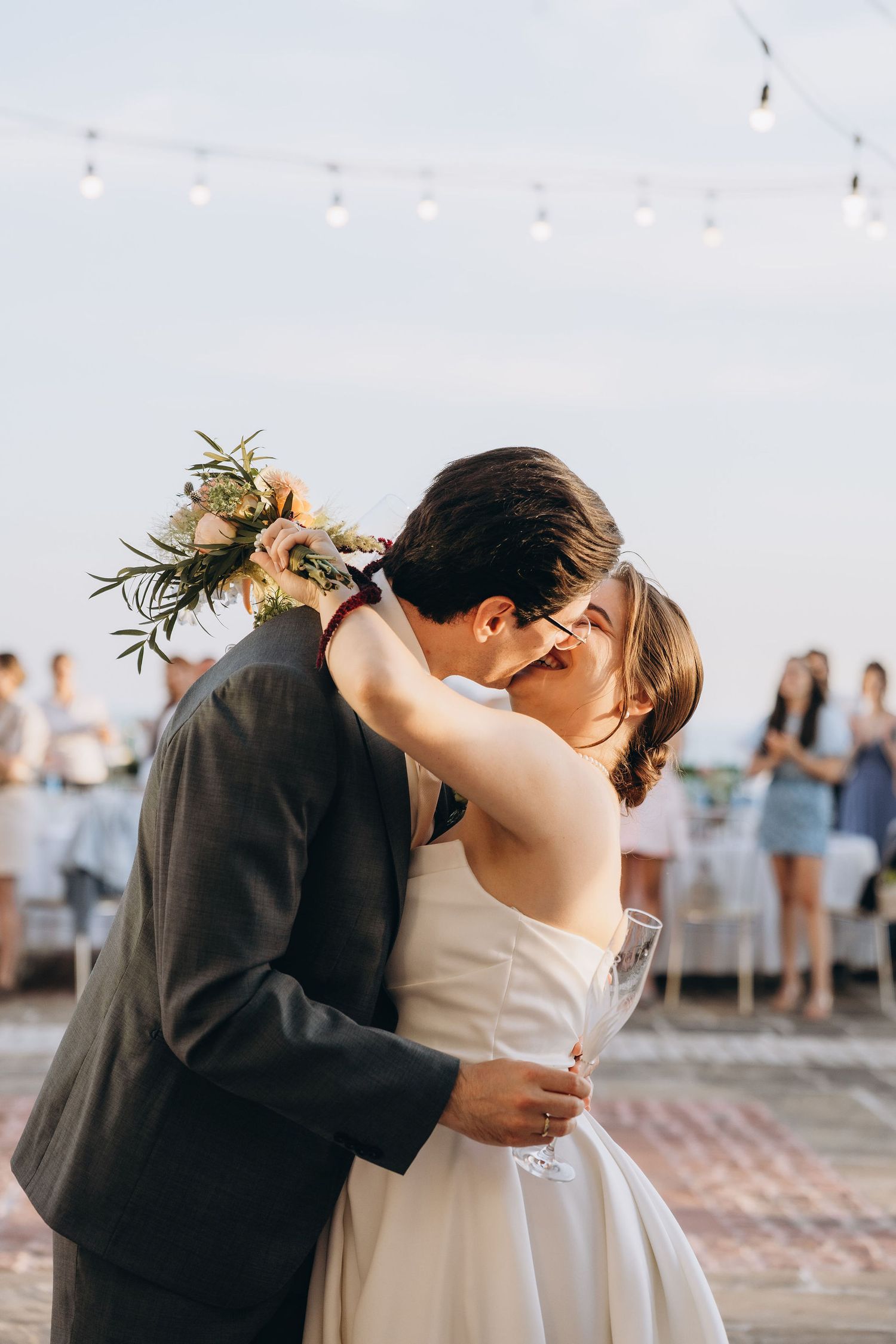 Bride and groom share first dance kiss at outdoor wedding reception with string lights and bouquet.