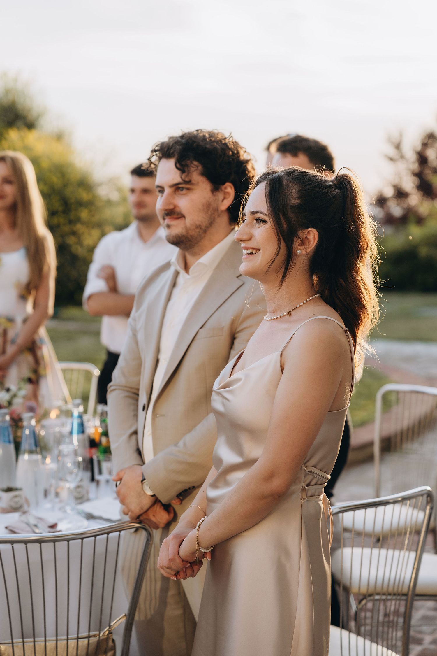 Couple in elegant attire smiling at outdoor wedding reception during golden hour.