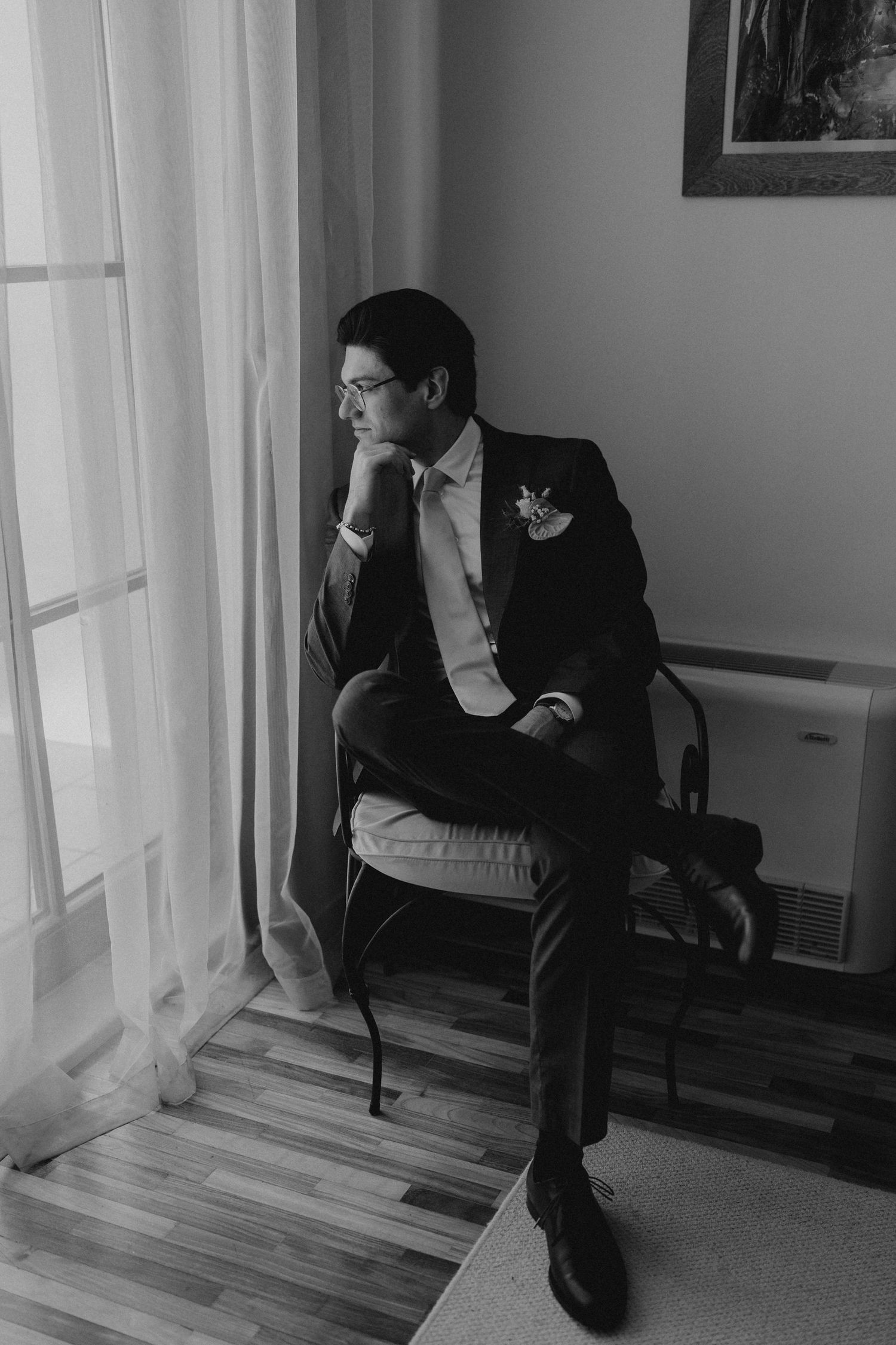 A groom in a suit sits pensively by a window, resting his chin on his hand, in a black and white wedding photo.