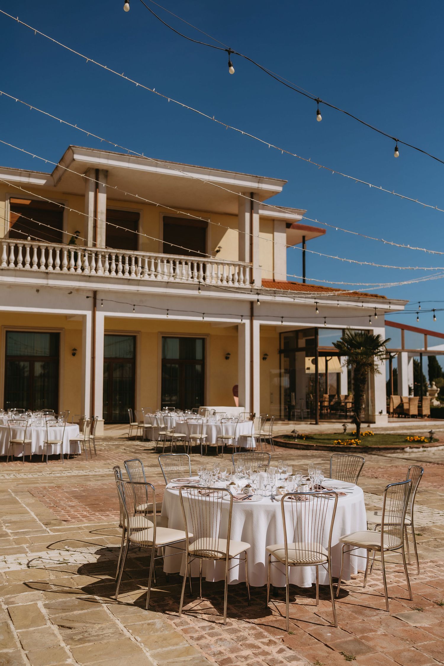 Elegant outdoor wedding reception setup with round white-clothed tables, string lights, and a classic villa backdrop.