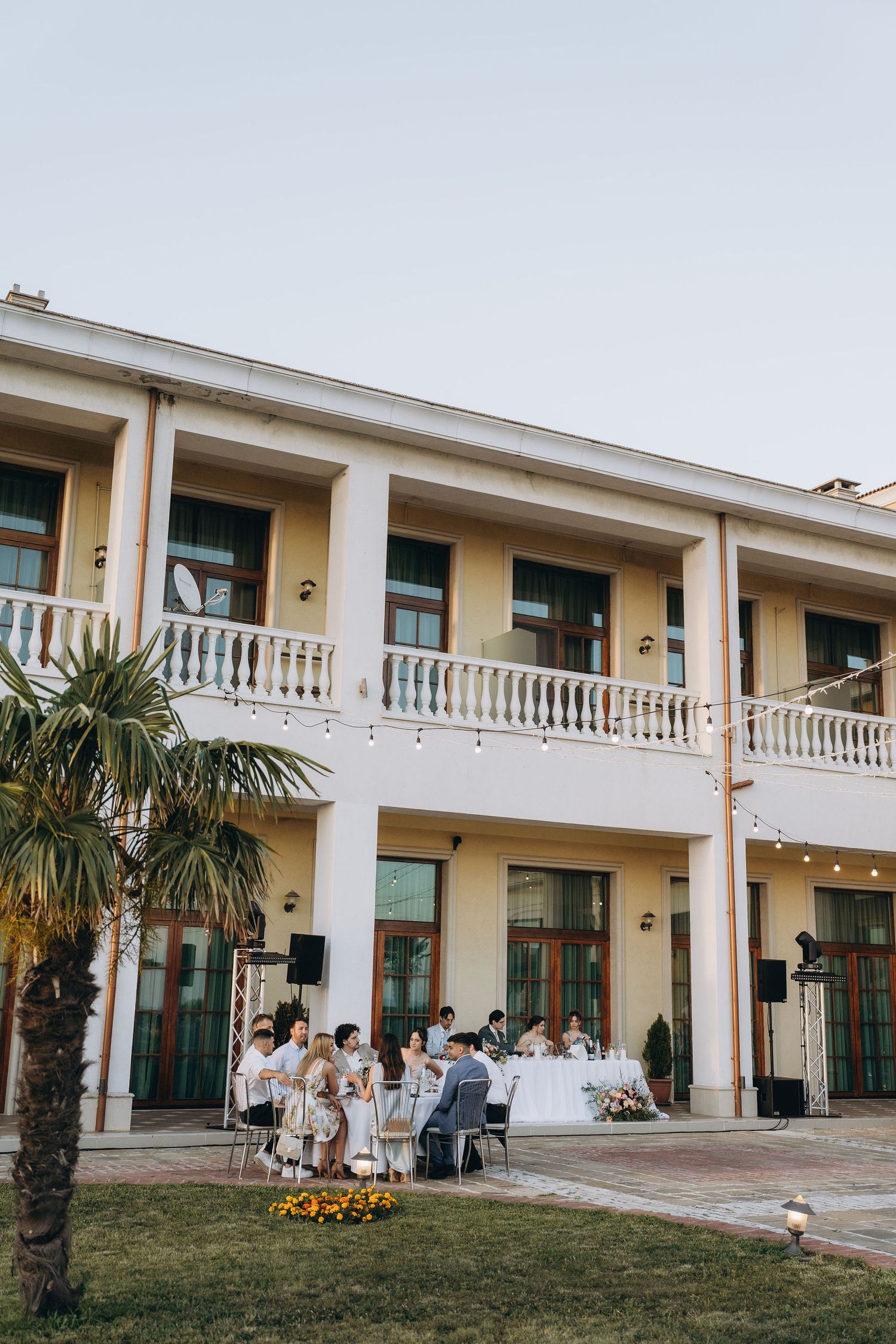 Elegant outdoor wedding reception on a patio of a two-story colonial building with white balcony and string lights.