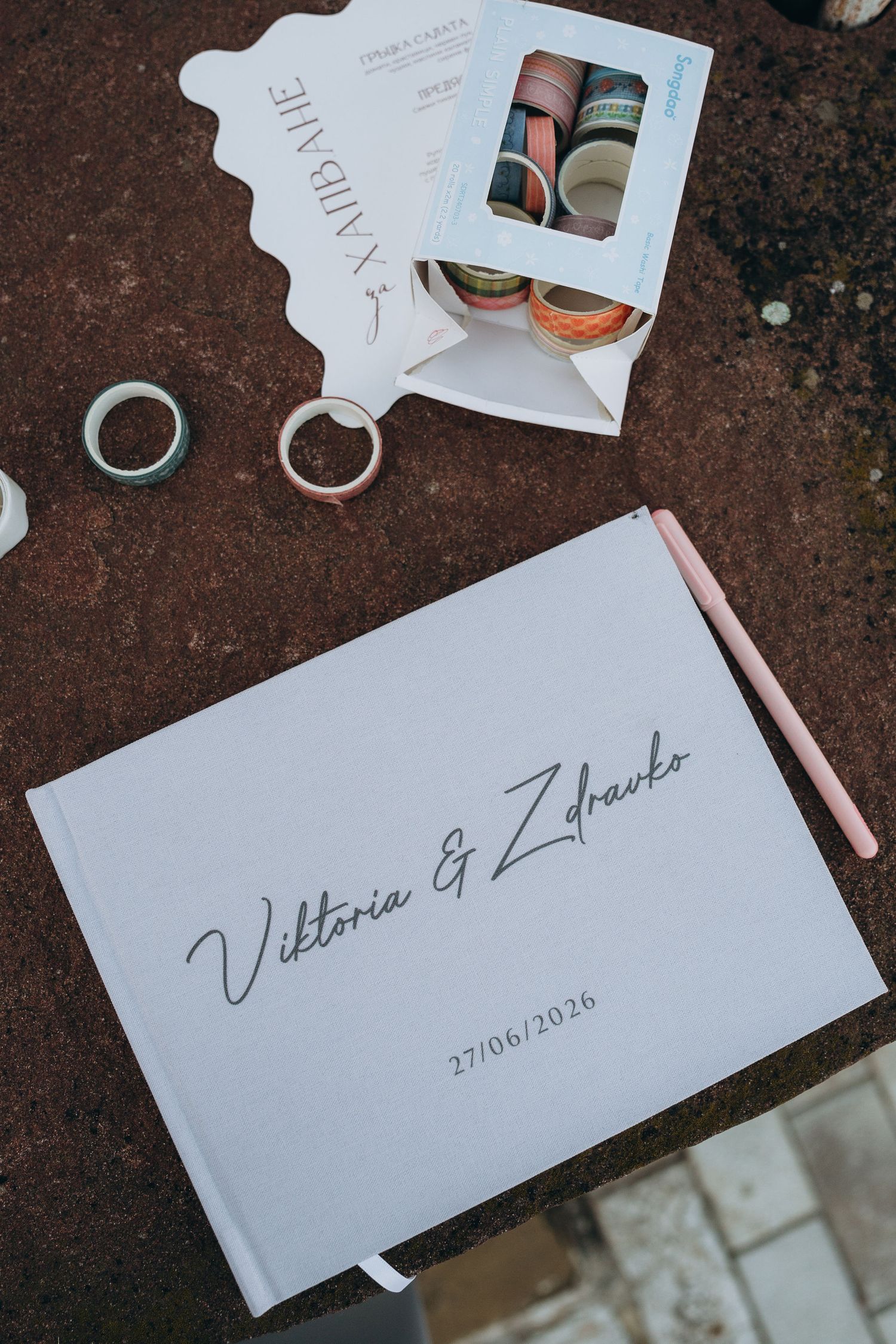 White wedding guest book for Viktoria and Zdravko dated 27/06/2026, with a pink pen, rings, and washi tape nearby.