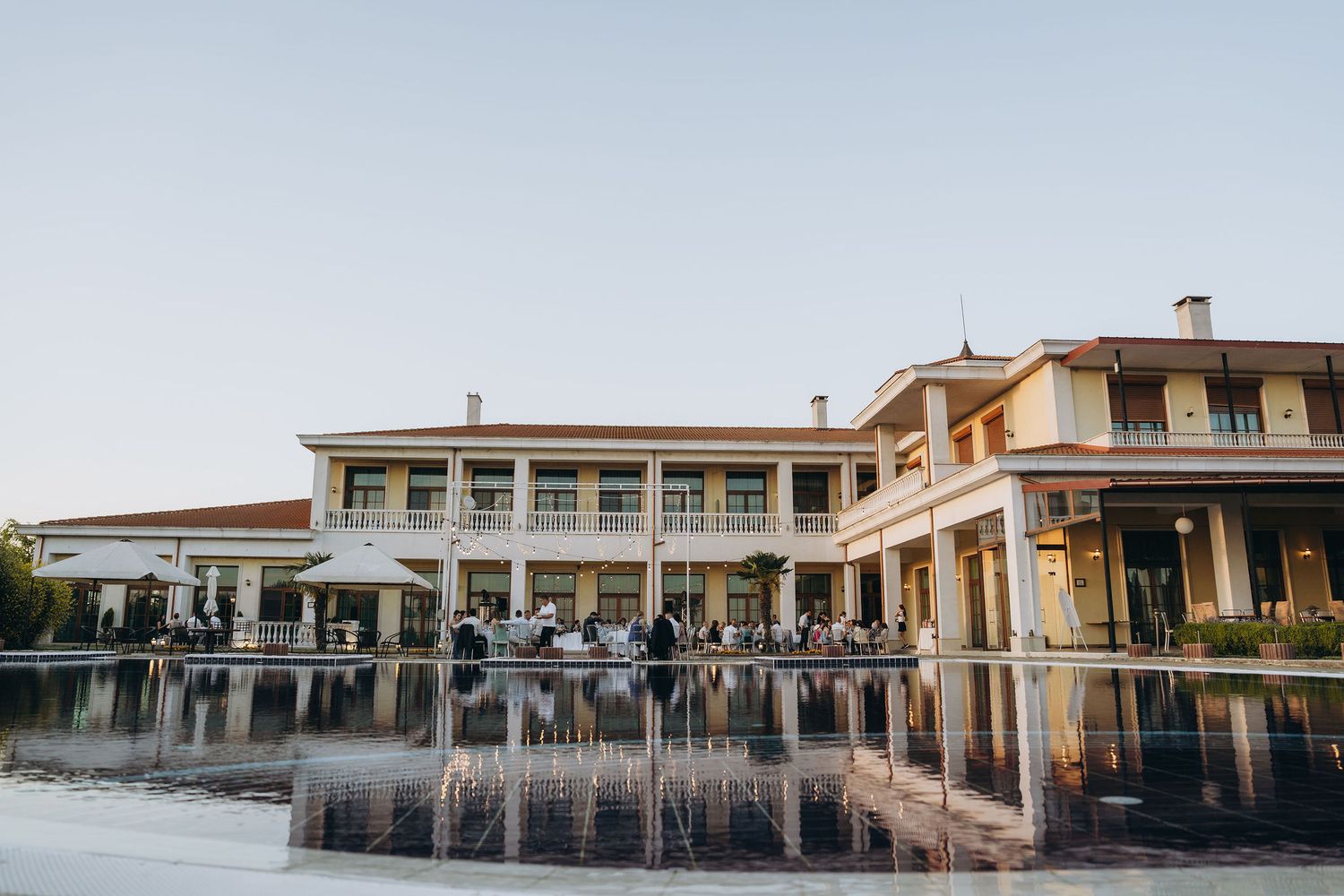 Elegant two-story venue with reflective pool at dusk, guests gathered on terrace for outdoor event.