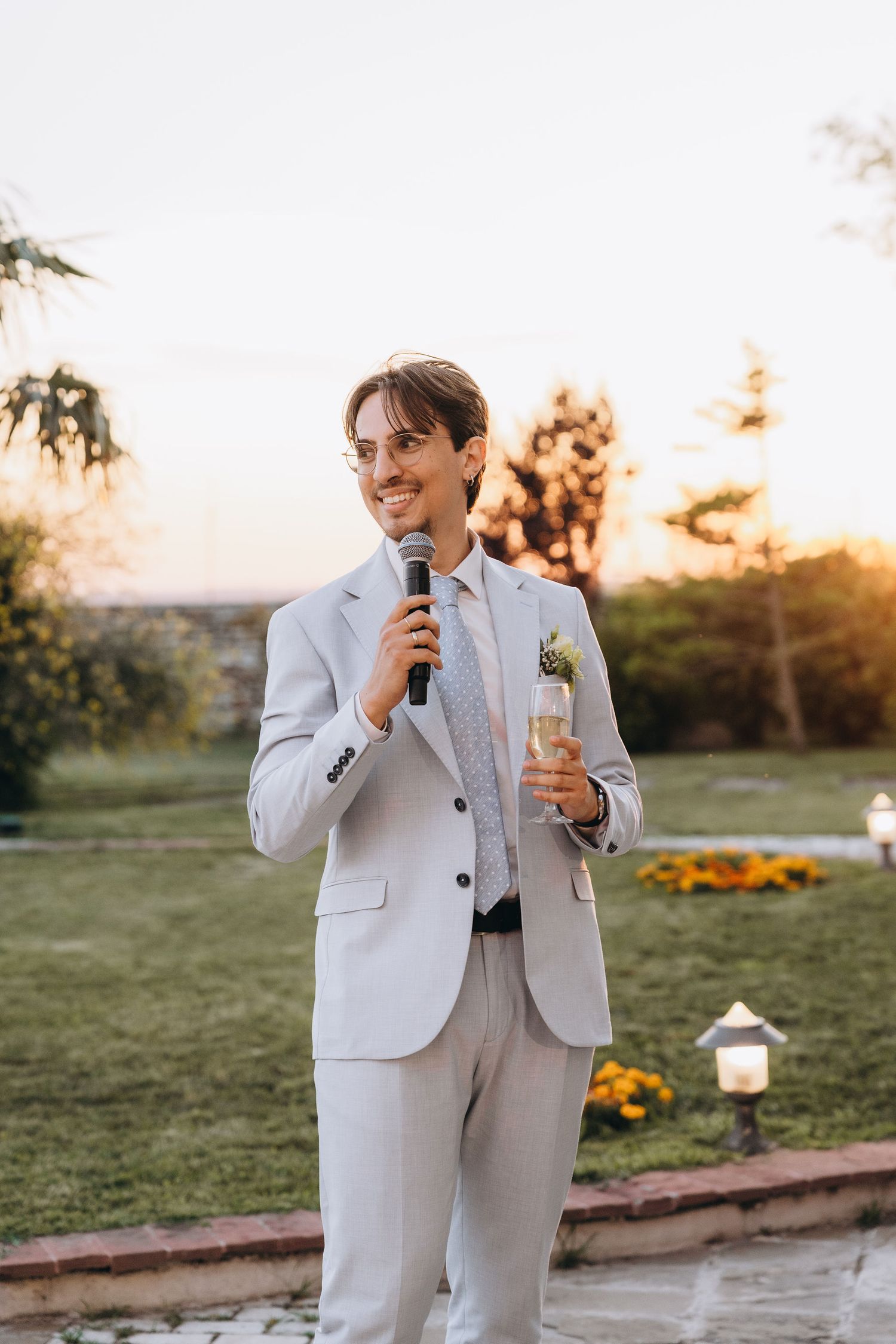 Smiling groom in light gray suit holds microphone and champagne glass while giving outdoor wedding speech at sunset.