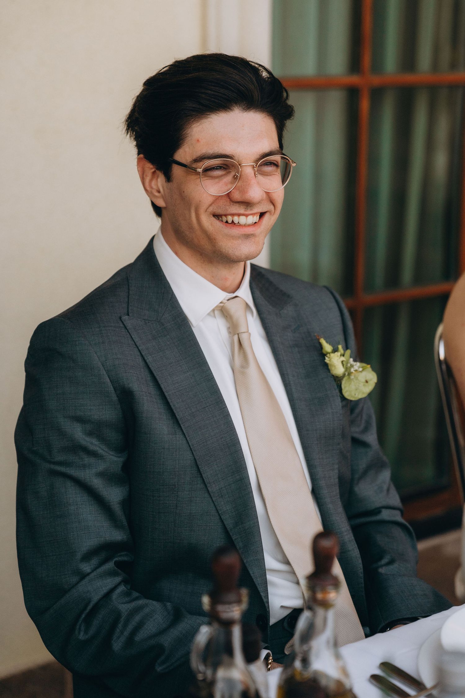 Smiling groom in gray suit with boutonniere and cream tie at wedding reception.