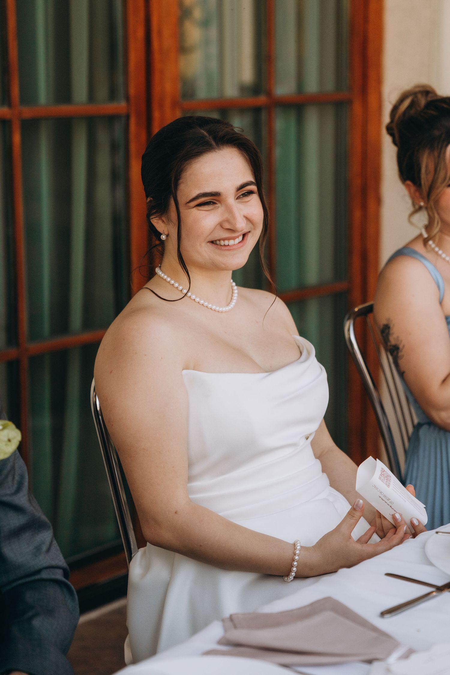 Smiling bride in white strapless gown and pearl jewelry seated at wedding reception table.