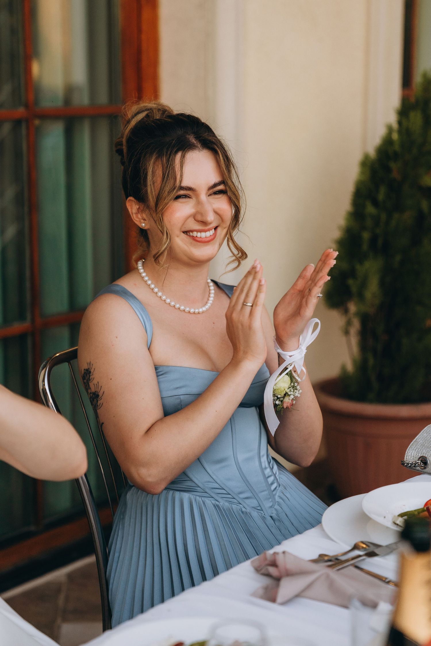 Smiling bridesmaid in blue dress and pearl necklace clapping at outdoor wedding reception table.