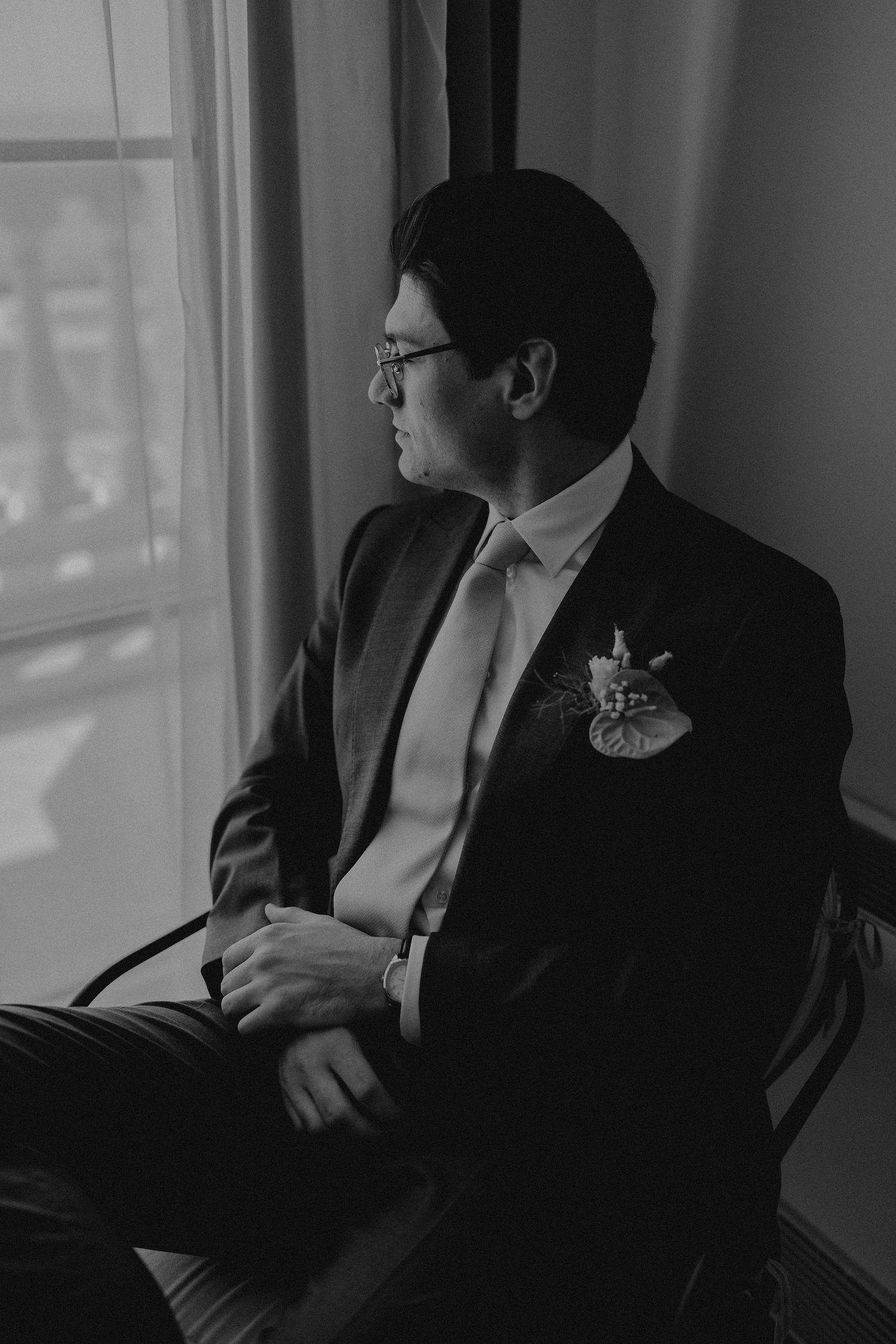 Black and white portrait of a groom in a suit with boutonniere, seated by a window, gazing thoughtfully to the side.