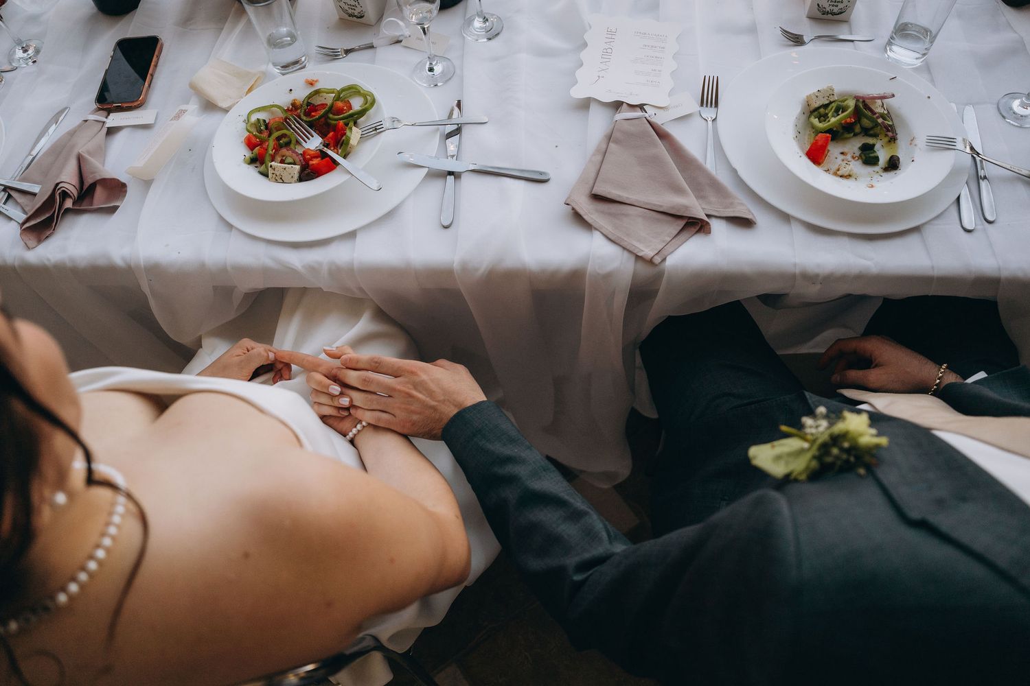 Bride and groom holding hands at wedding reception table with elegant place settings and fresh salads.