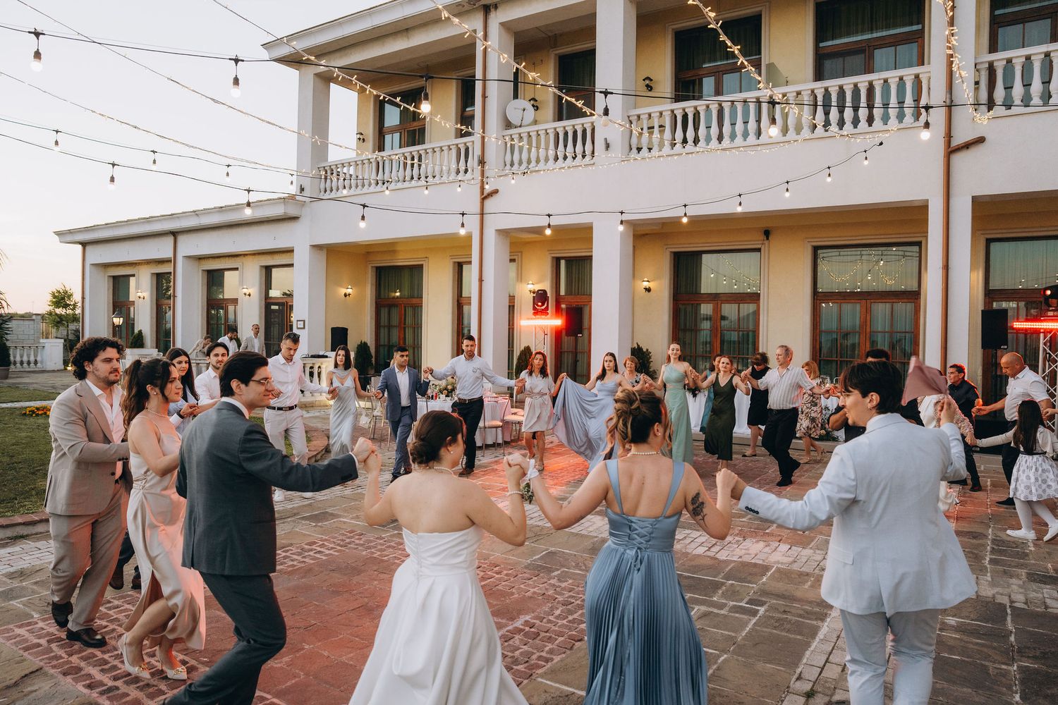 Wedding guests dancing in a circle outdoors at an elegant venue with string lights and a grand white building backdrop.