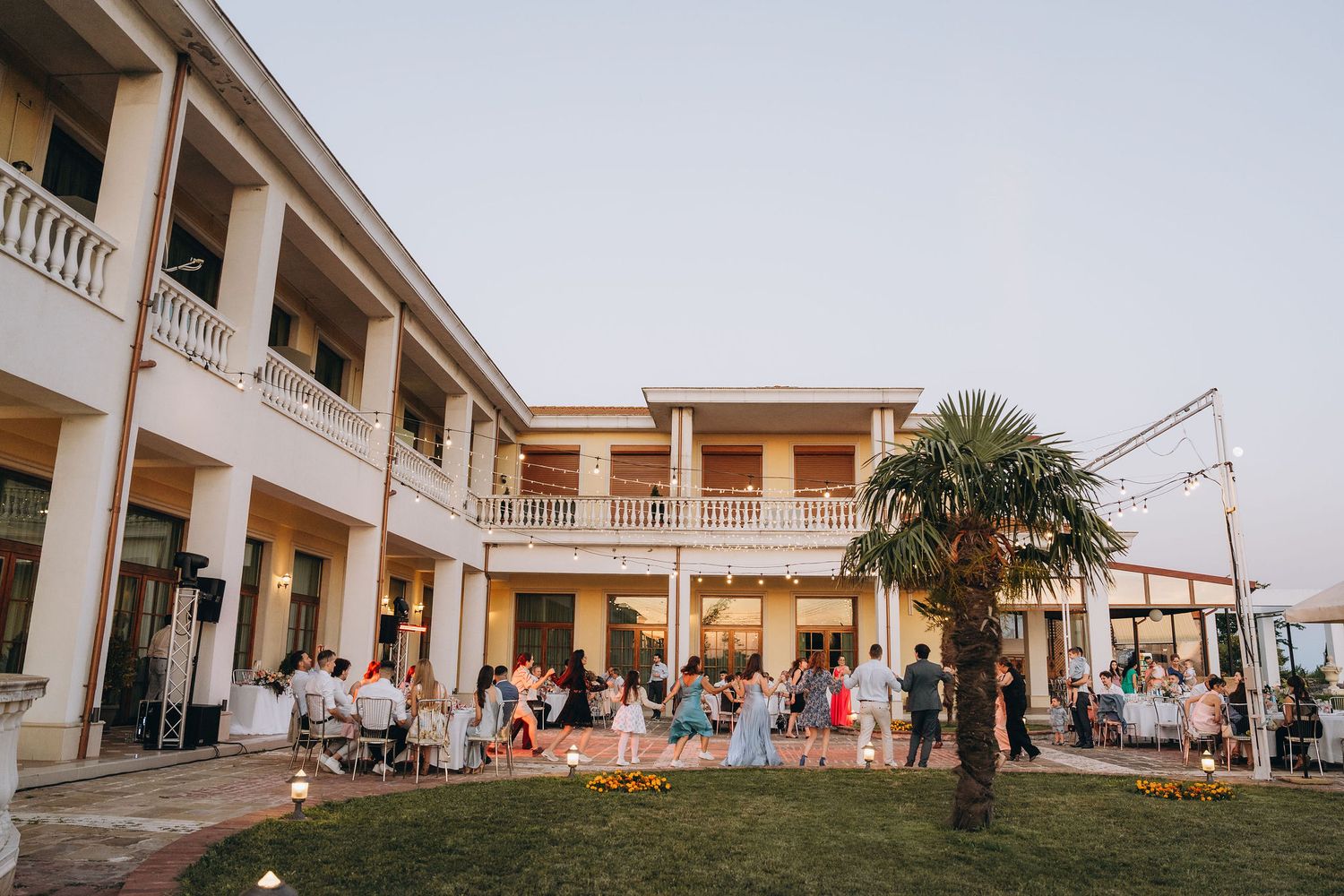 Elegant two-story white venue with guests gathered on lawn at dusk, featuring balconies and palm tree.