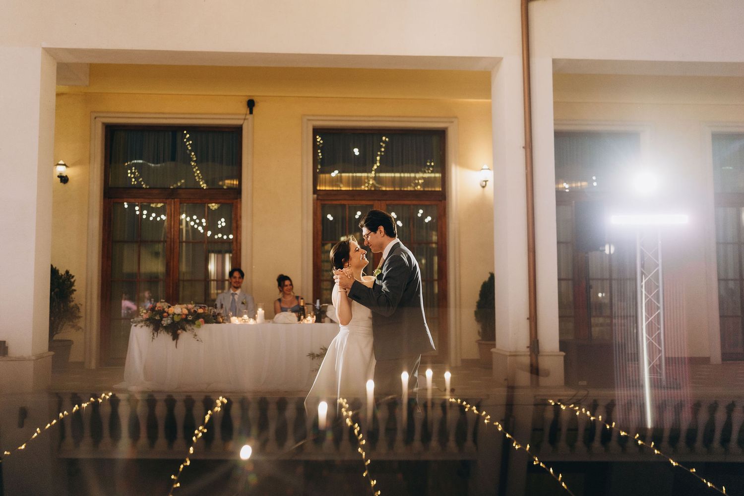 Bride and groom share first dance on romantic candlelit outdoor terrace adorned with fairy lights at evening wedding reception.