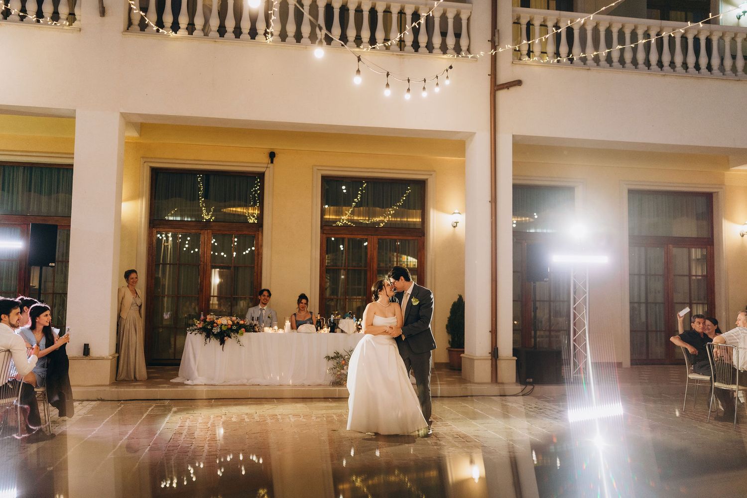 Bride and groom share first dance in elegant ballroom with string lights and guests watching at wedding reception.