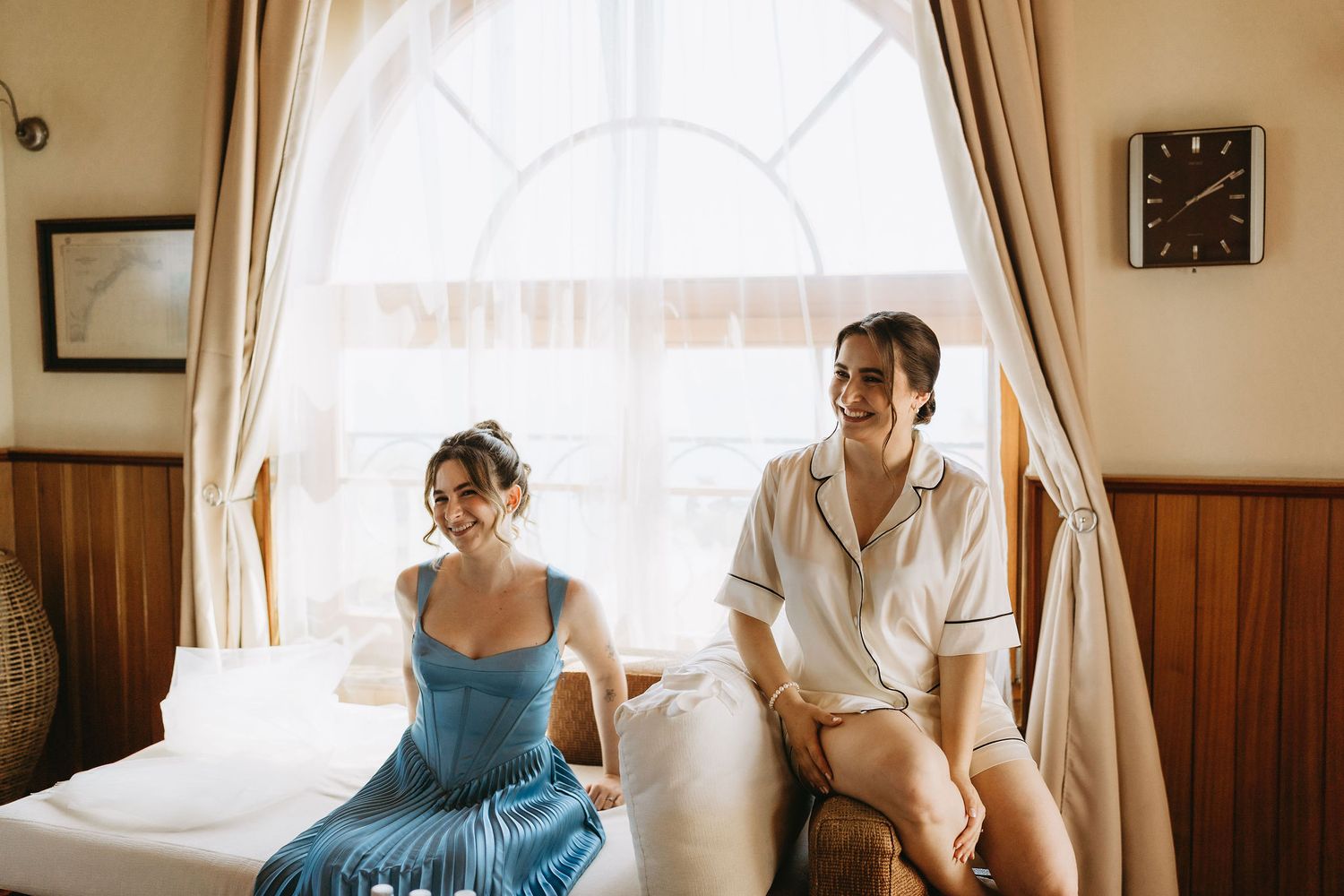 Two smiling women sitting on a bed by a bright window in a cozy wooden room, one in a blue dress.