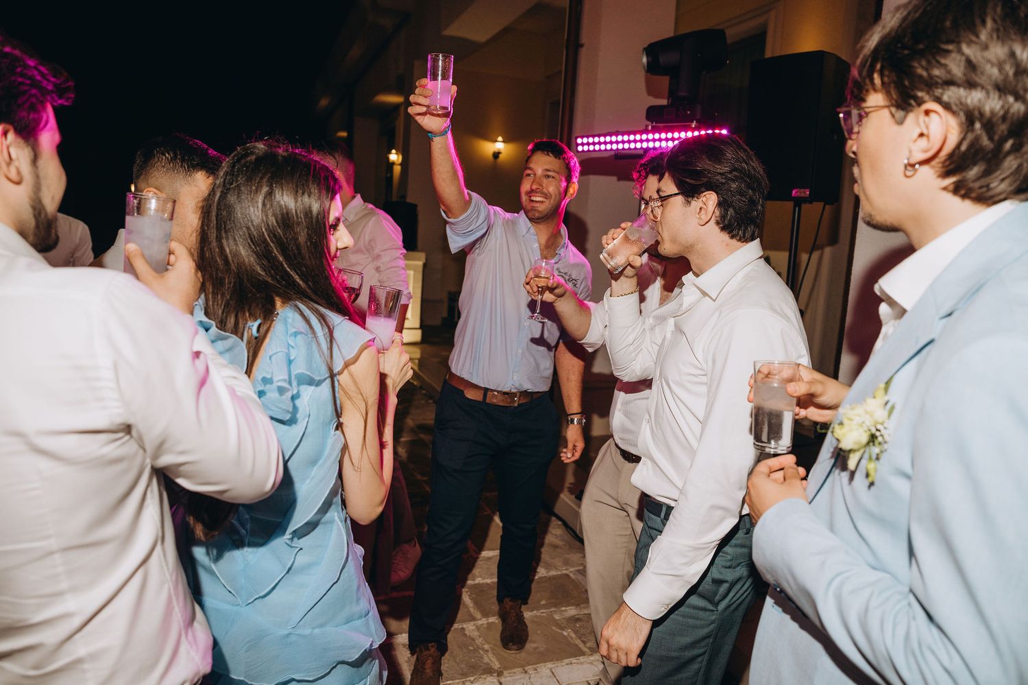 Group of young adults dancing and celebrating at an outdoor evening party, raising drinks in a festive atmosphere.