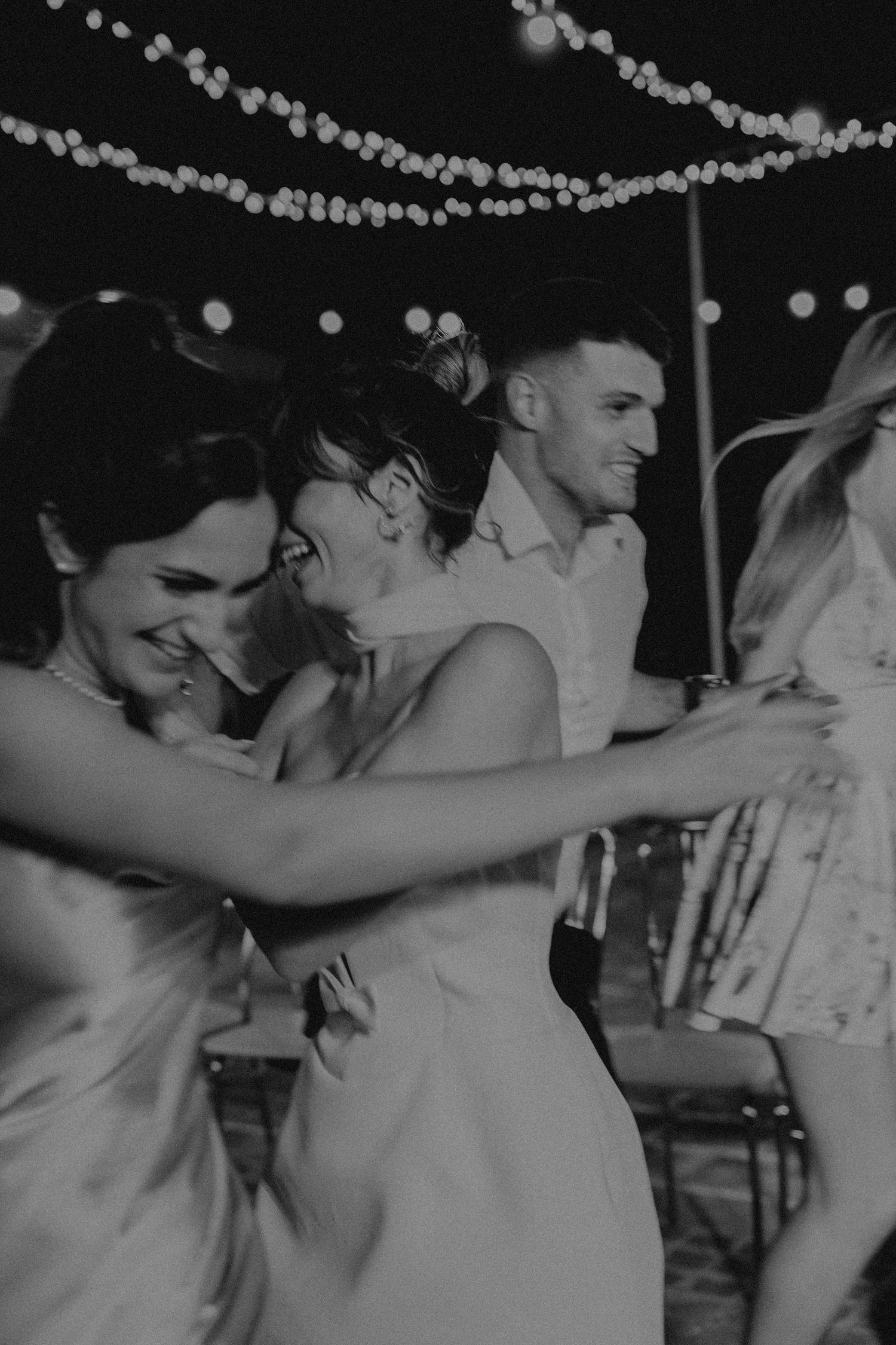 Black and white photo of guests laughing and dancing together at an outdoor wedding reception with string lights overhead.