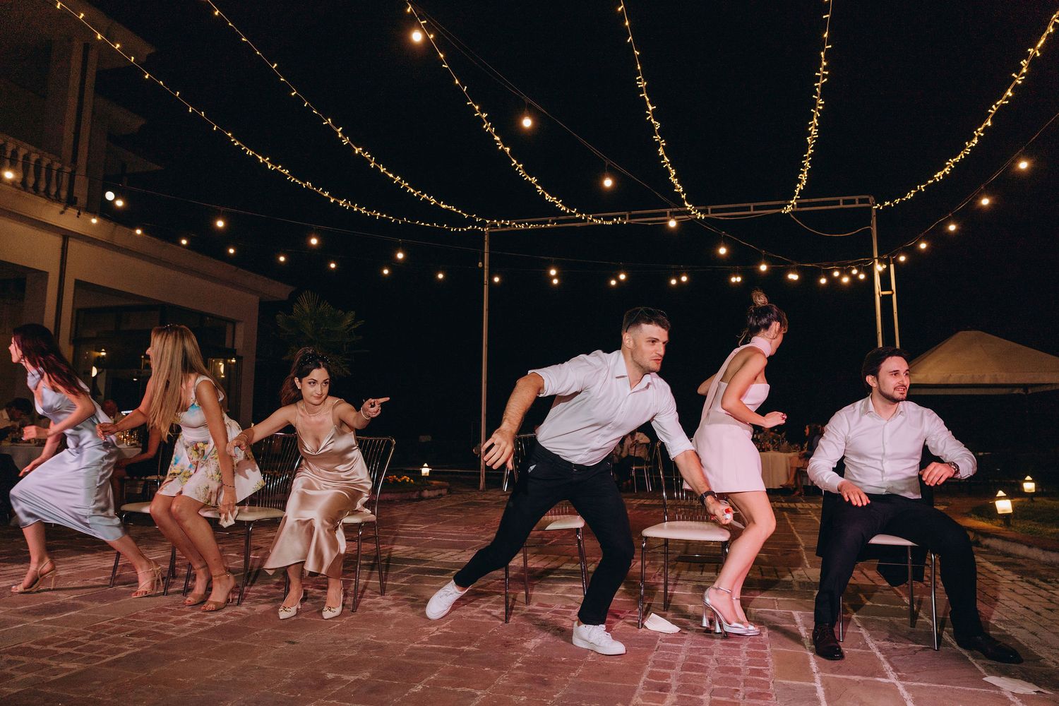 Group of guests dancing outdoors at night under string lights at a festive wedding reception party.