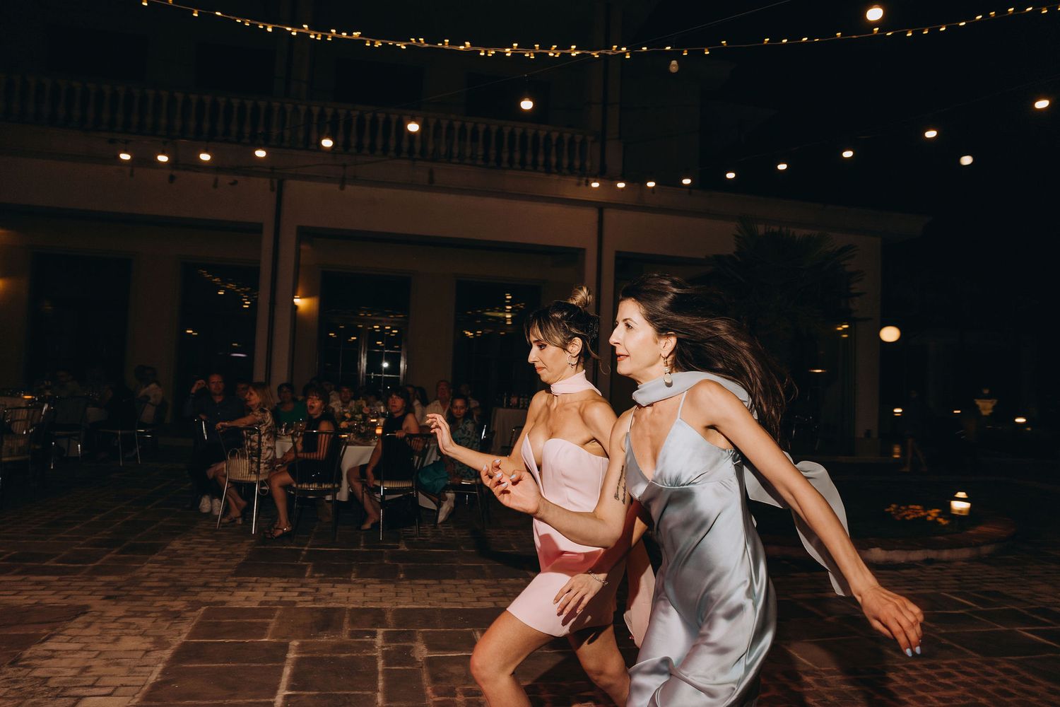 Two women in elegant dresses dancing joyfully at an outdoor evening wedding reception with string lights.