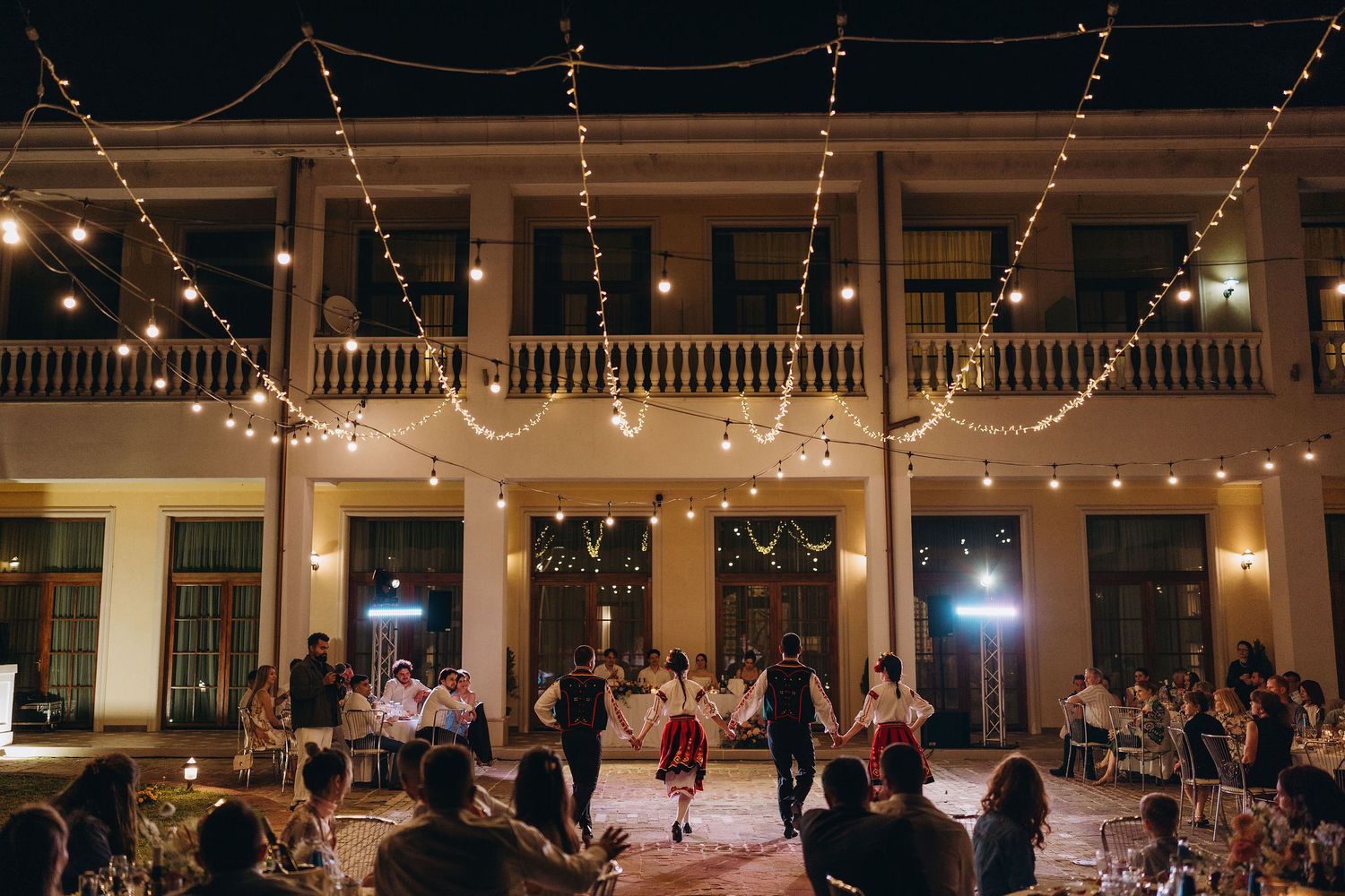 Outdoor evening wedding reception with string lights and folk dancers performing in a courtyard of an elegant venue.