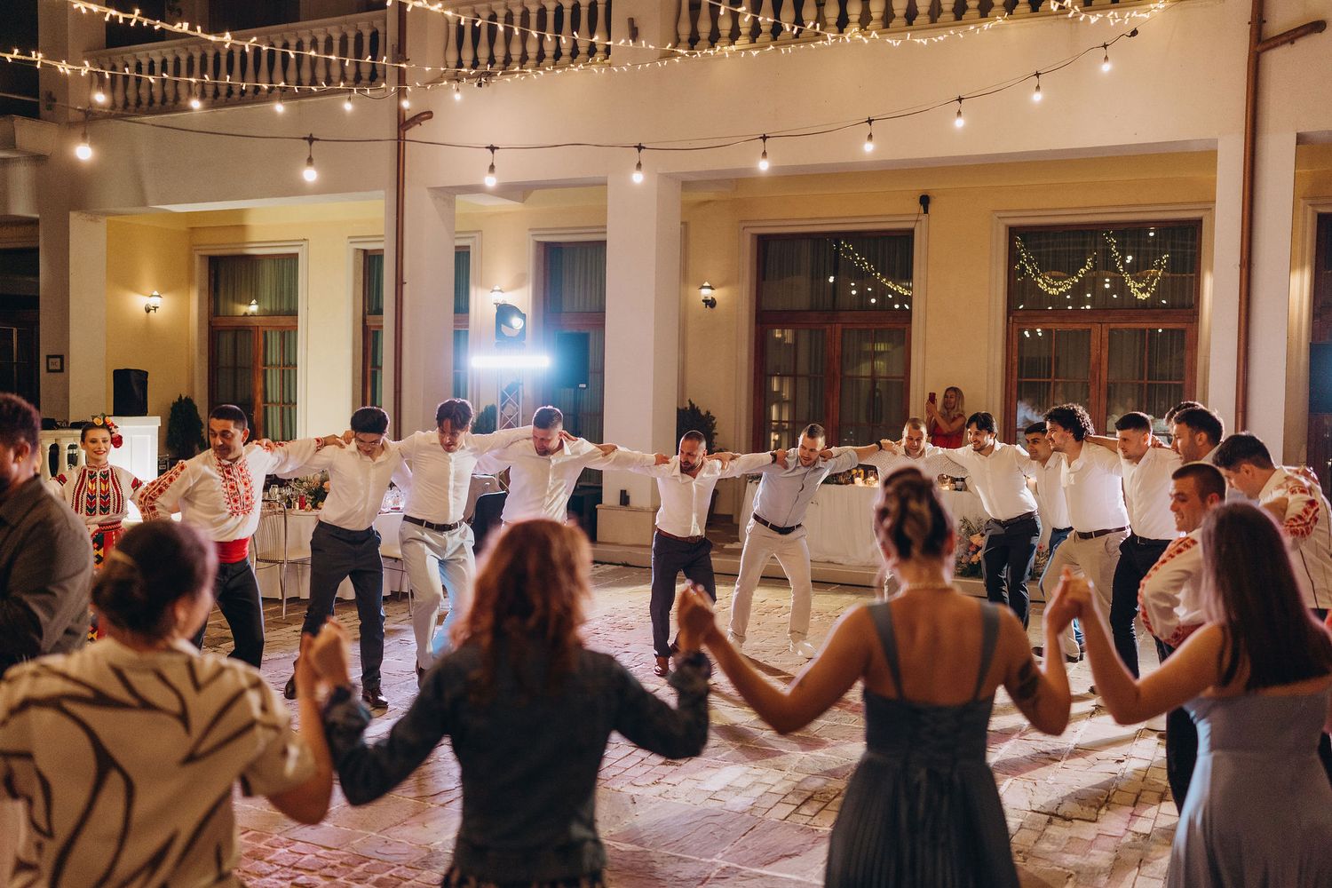 Wedding guests holding hands in a circle dance on a decorated ballroom floor with string lights overhead.