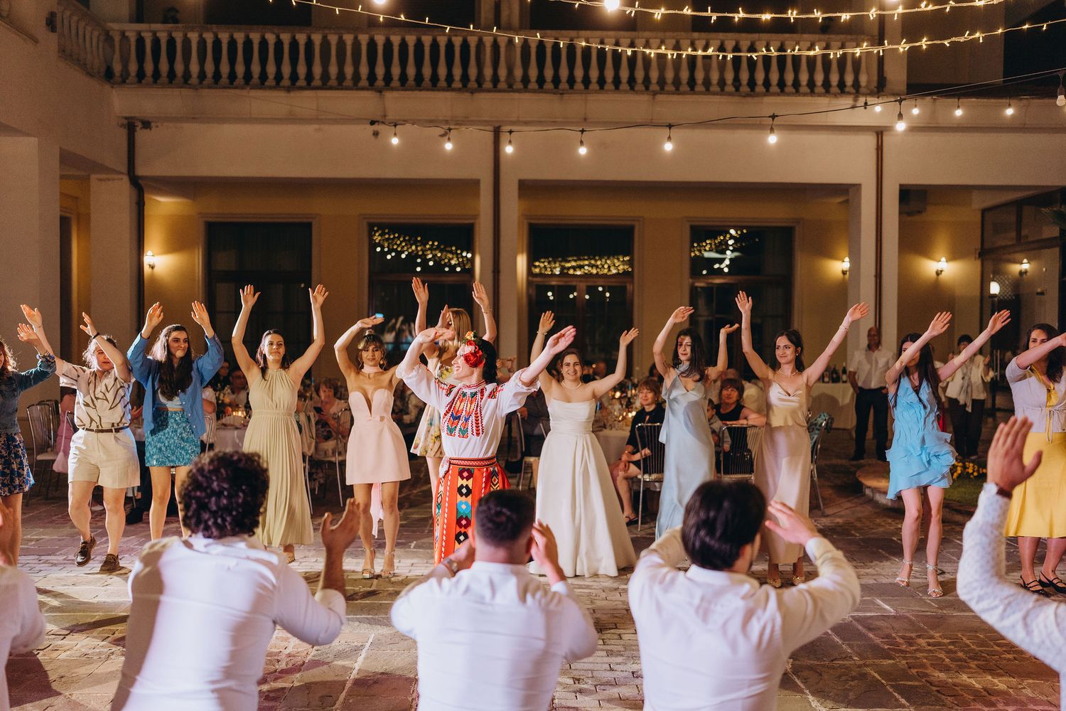 Wedding guests dancing with arms raised at elegant venue with string lights during lively reception celebration.