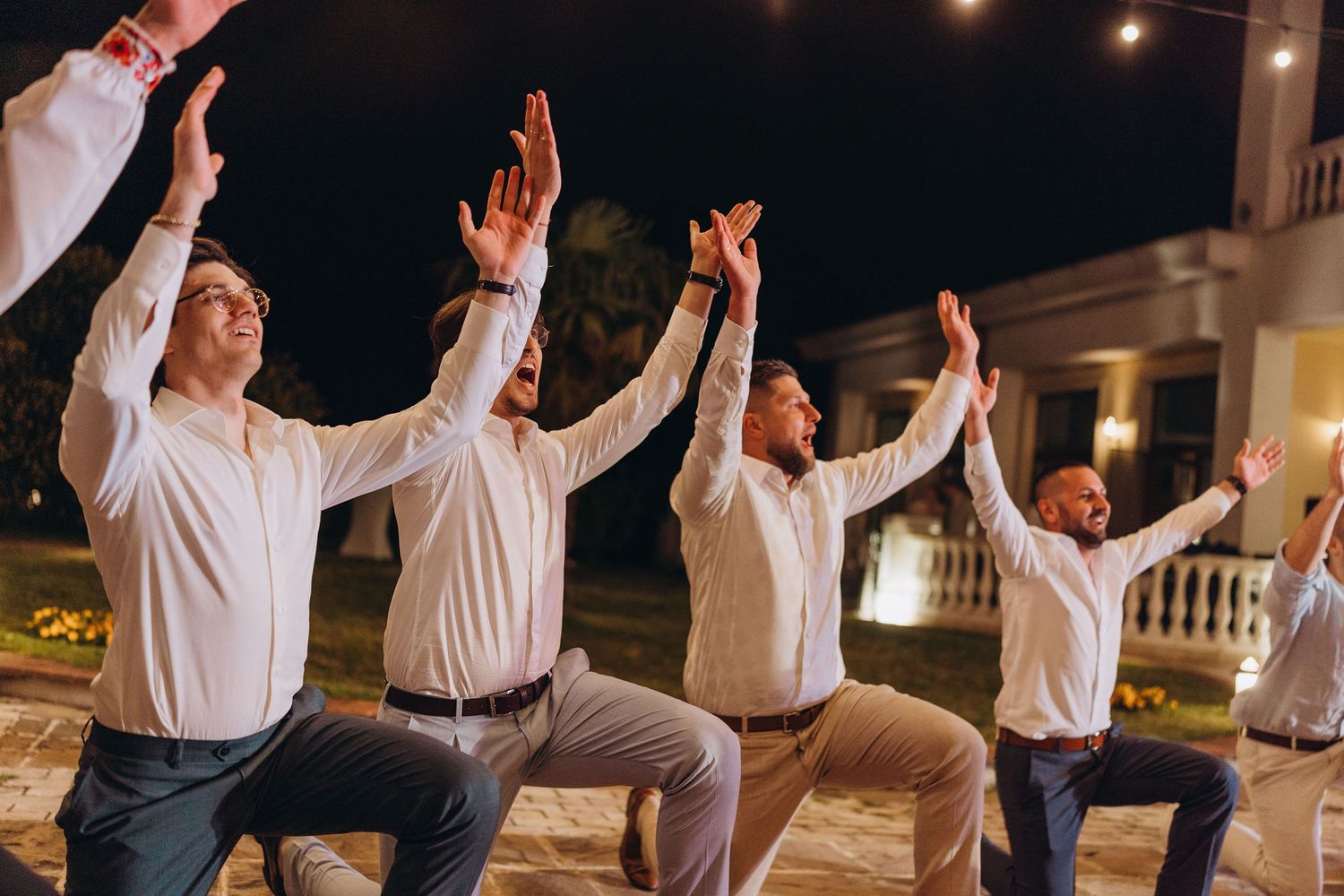 Four men in white shirts performing a synchronized dance move outdoors at night during a wedding celebration.