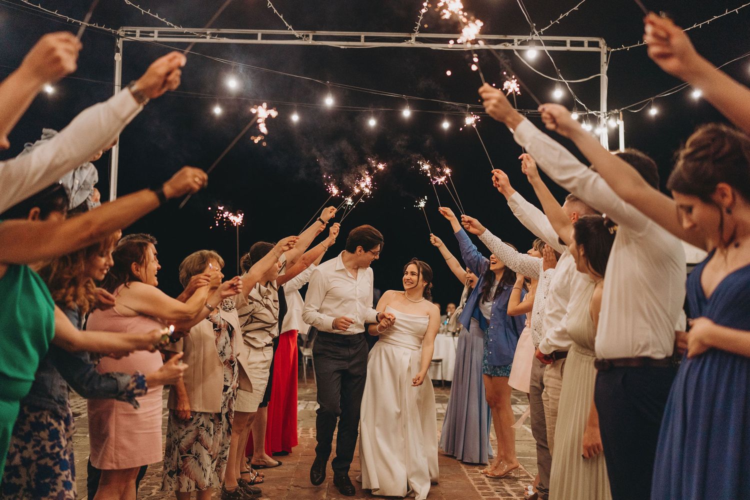 Bride and groom walk through sparkler tunnel surrounded by wedding guests at night outdoor reception.