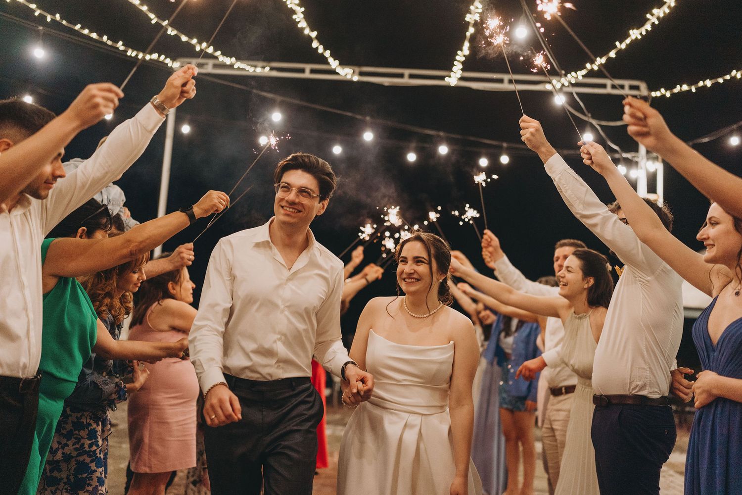 Bride and groom walk through sparkler tunnel surrounded by cheering guests at romantic outdoor wedding reception.