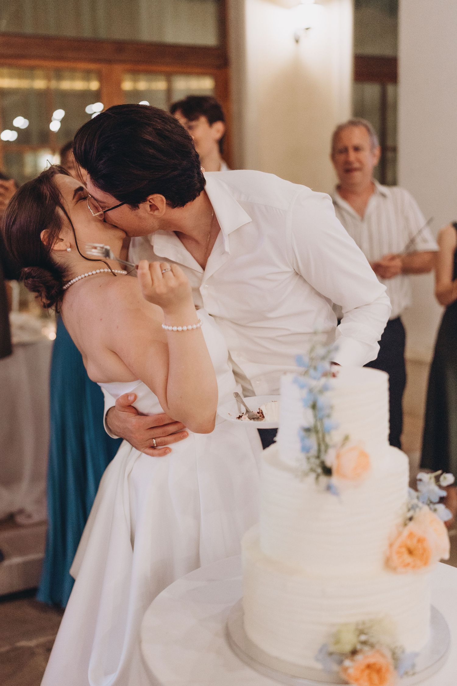 Bride and groom sharing a kiss beside a white three-tier wedding cake decorated with pastel flowers at their reception.