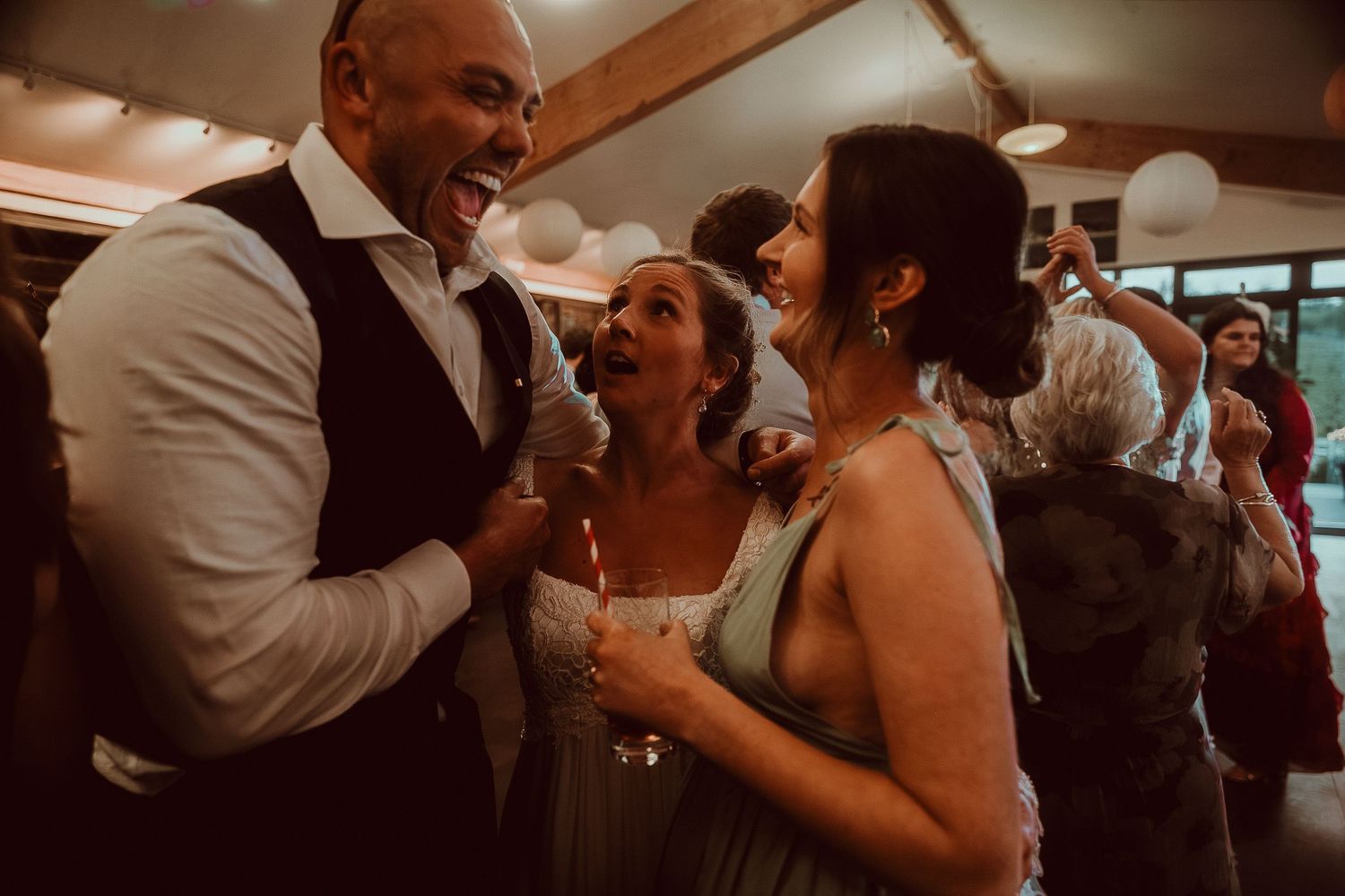 Joyful wedding reception guests laughing and dancing together in a warmly lit venue with balloon decorations.