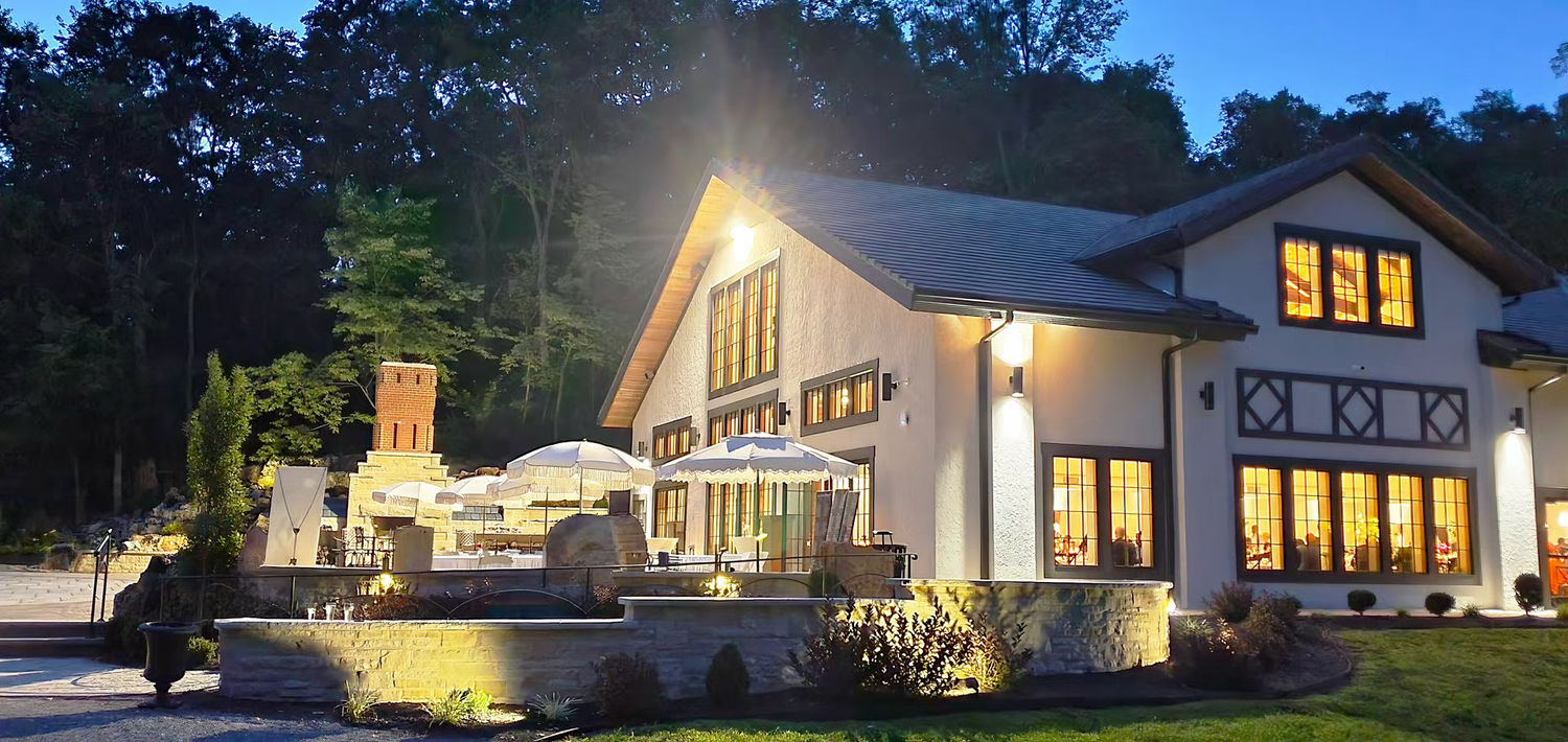 Elegant white building with warm glowing windows and outdoor patio illuminated at dusk surrounded by lush trees.