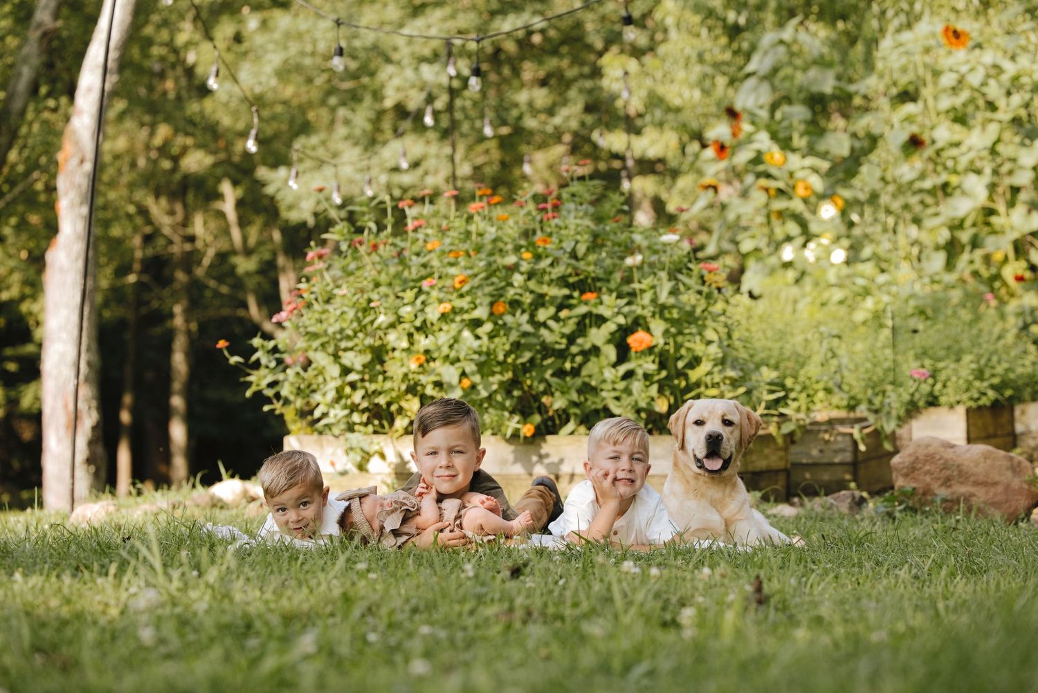 Four children and a golden retriever dog lying on green grass in a sunny garden with colorful flowers.