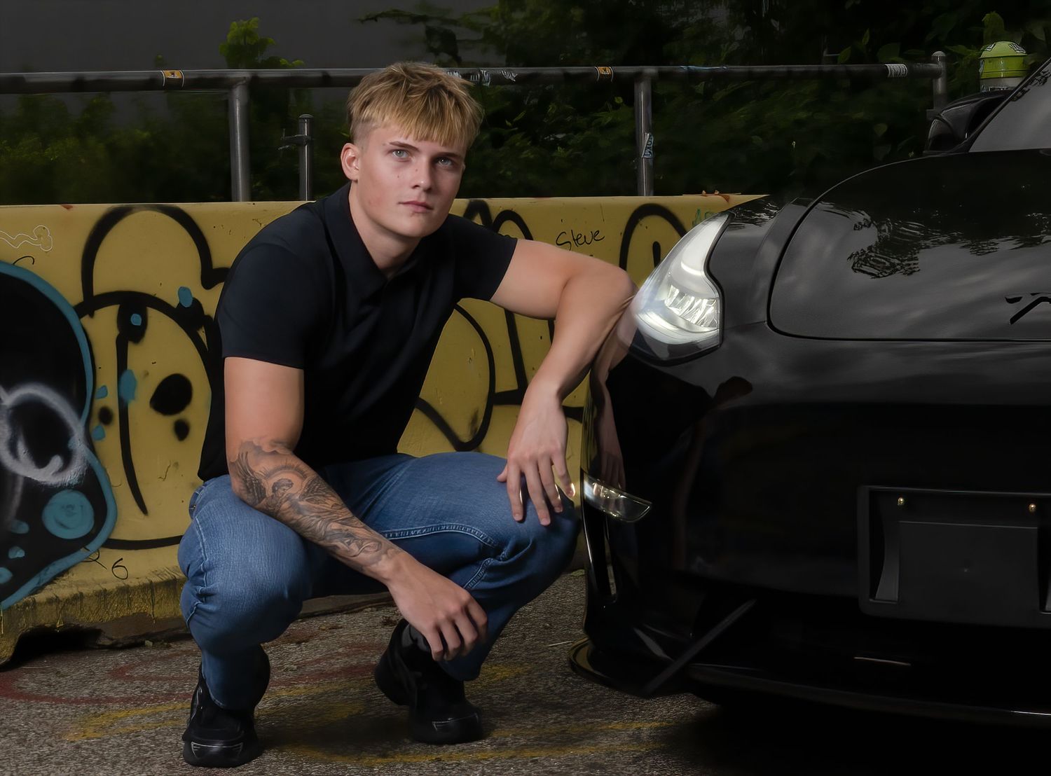Young tattooed man crouching beside a black sports car near graffiti wall, holding a tool outdoors.