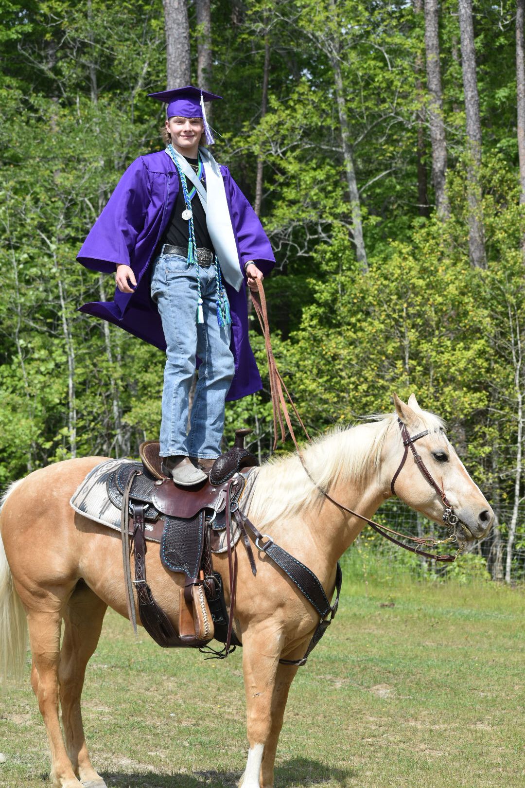 Graduate in purple cap and gown standing on palomino horse in grassy field.