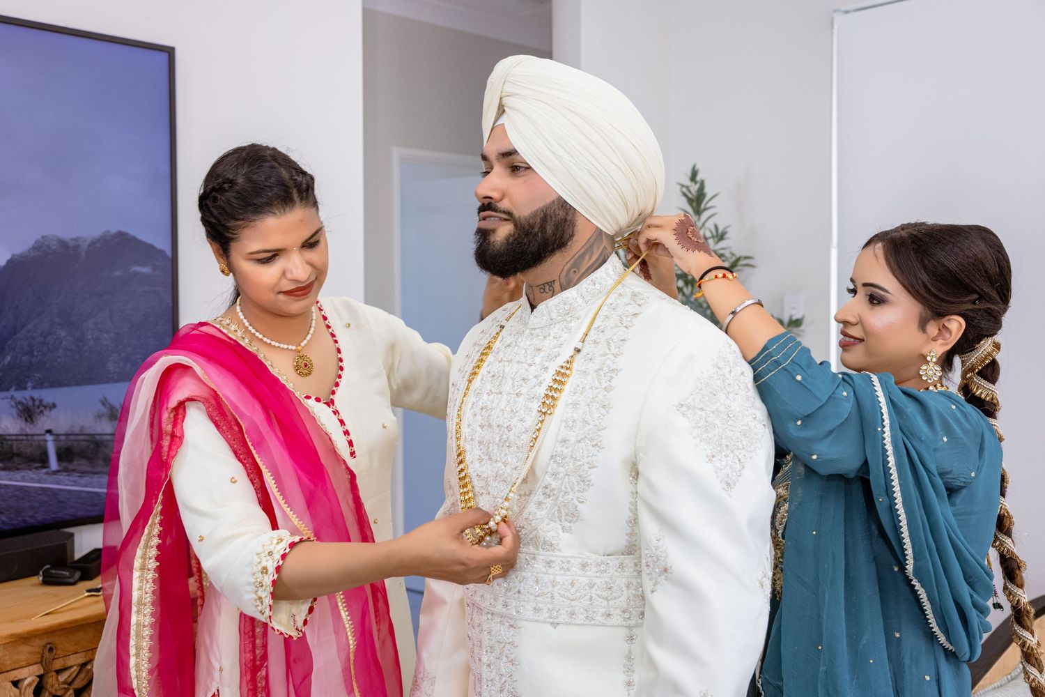 Two women help a Sikh groom in white sherwani and turban prepare for his wedding ceremony.