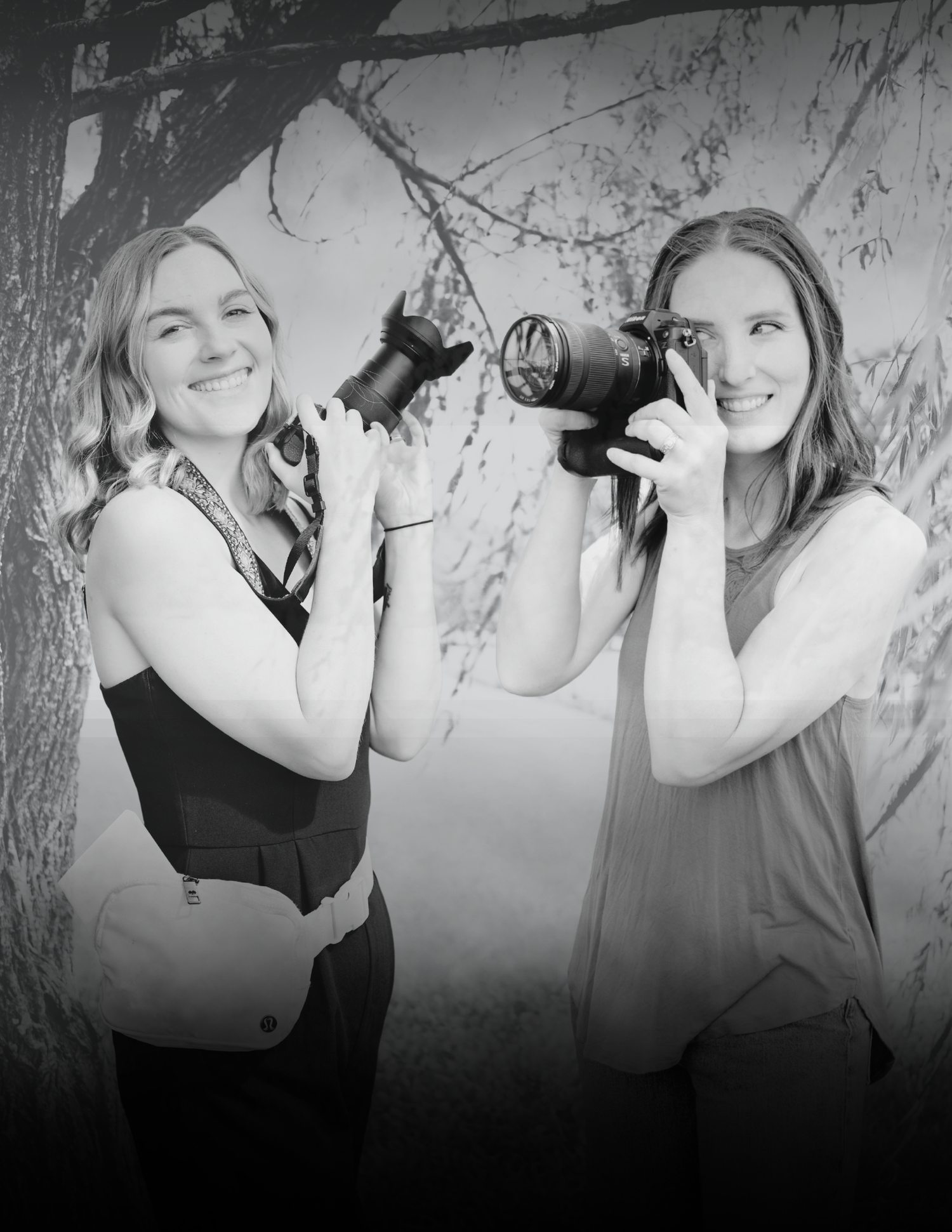 Two smiling women photographers holding cameras outdoors near trees in a black and white portrait photo.