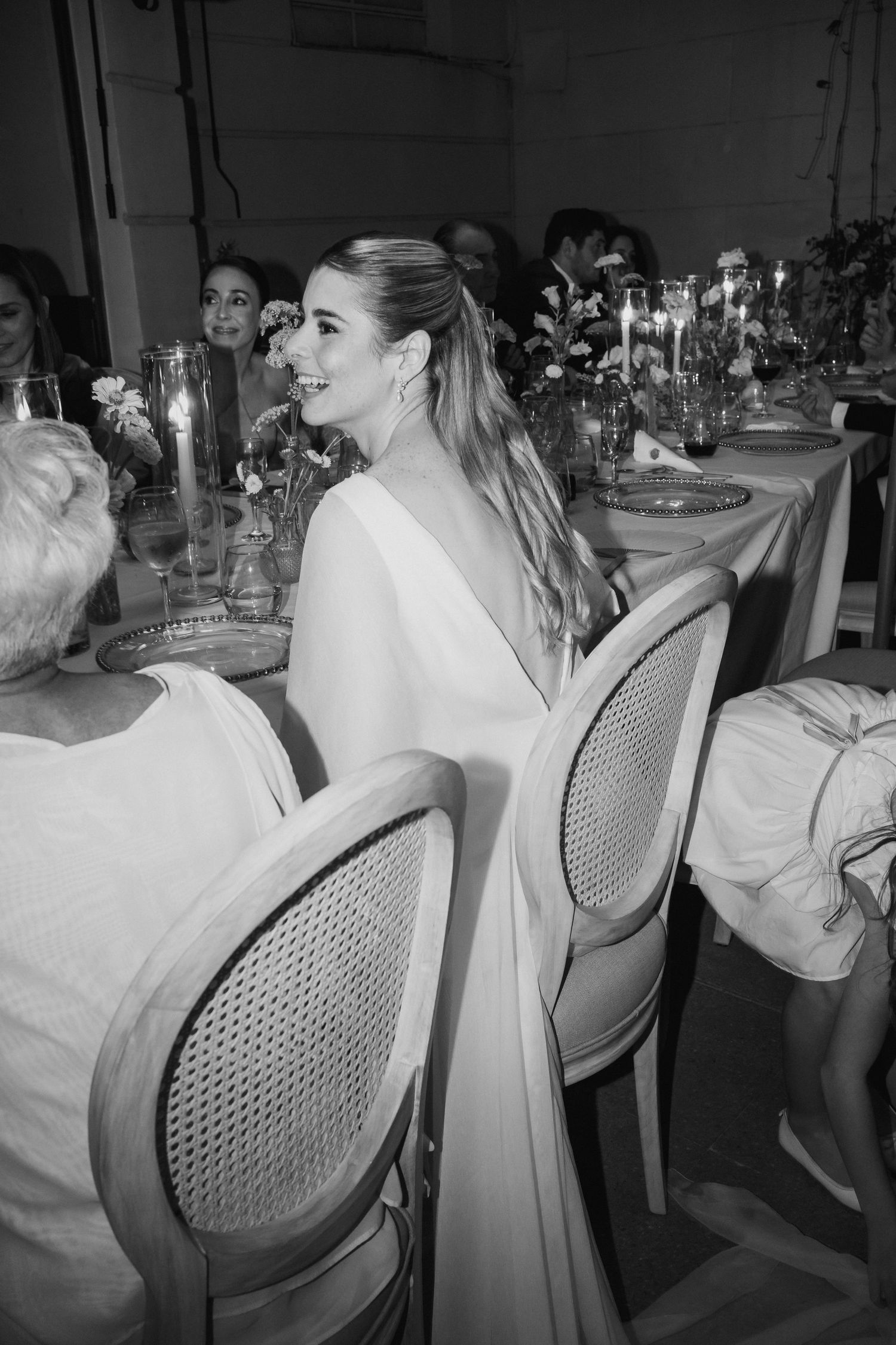 Smiling bride in white cape gown seated at elegant candlelit wedding reception table, black and white photo.