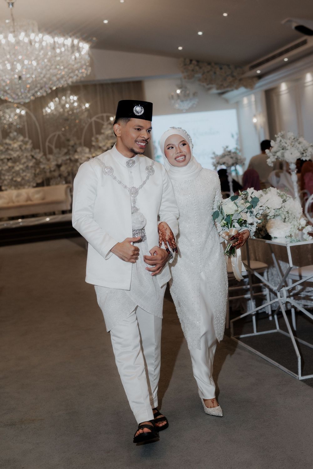 Muslim couple in white wedding attire walking together at elegant reception, groom in baju melayu, bride in hijab holding bouquet.