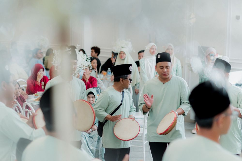 Men in traditional Malay attire playing rebana drums during a cultural ceremony indoors.