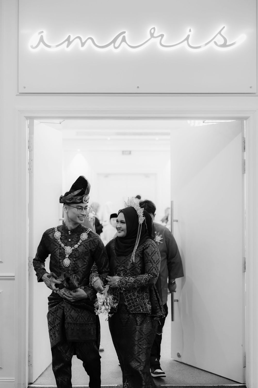 Malay couple in traditional attire walking through Amaris venue doorway in black and white photo.