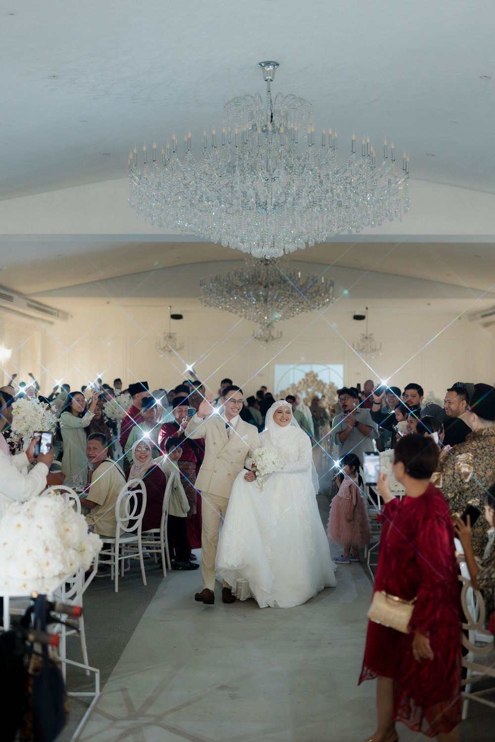 Bride and groom walk down the aisle at an elegant wedding reception with crystal chandeliers and guests cheering.