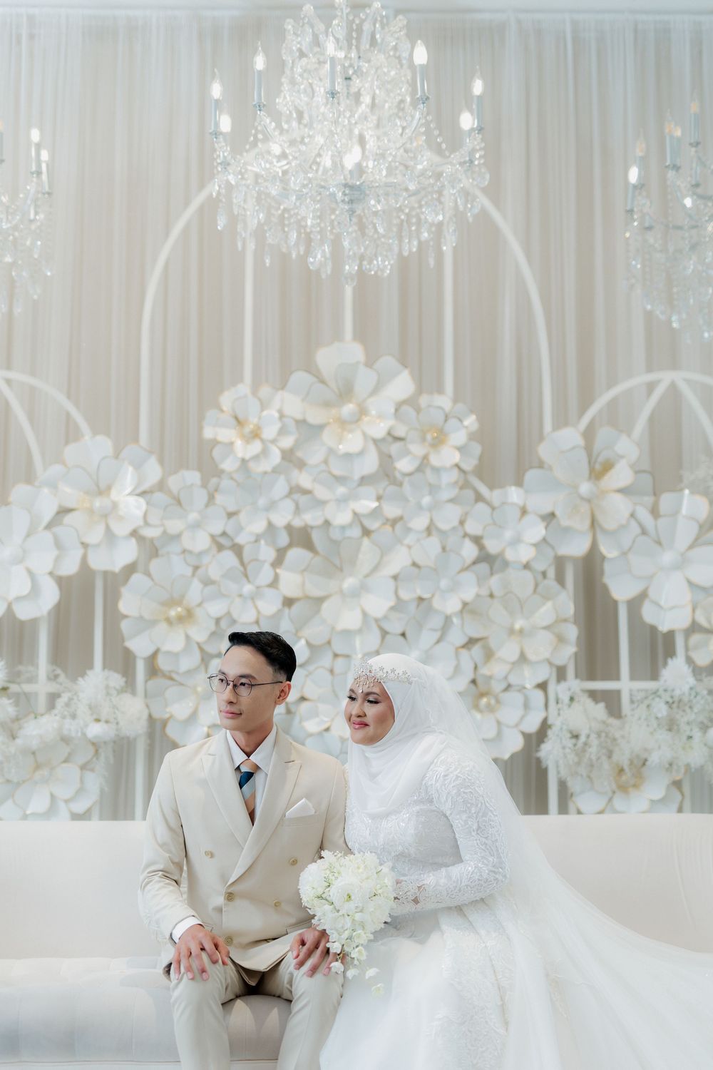Muslim bride and groom in white wedding attire seated before elegant floral backdrop with crystal chandelier.