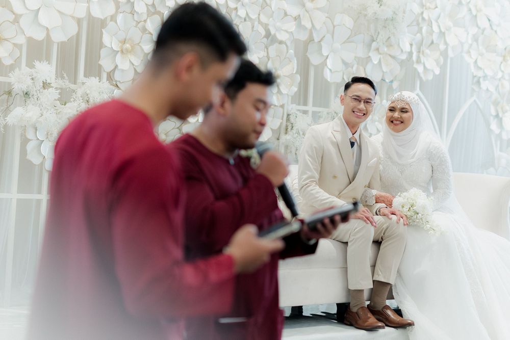 Muslim wedding ceremony with bride in hijab and groom in white suit seated while guests recite prayers.