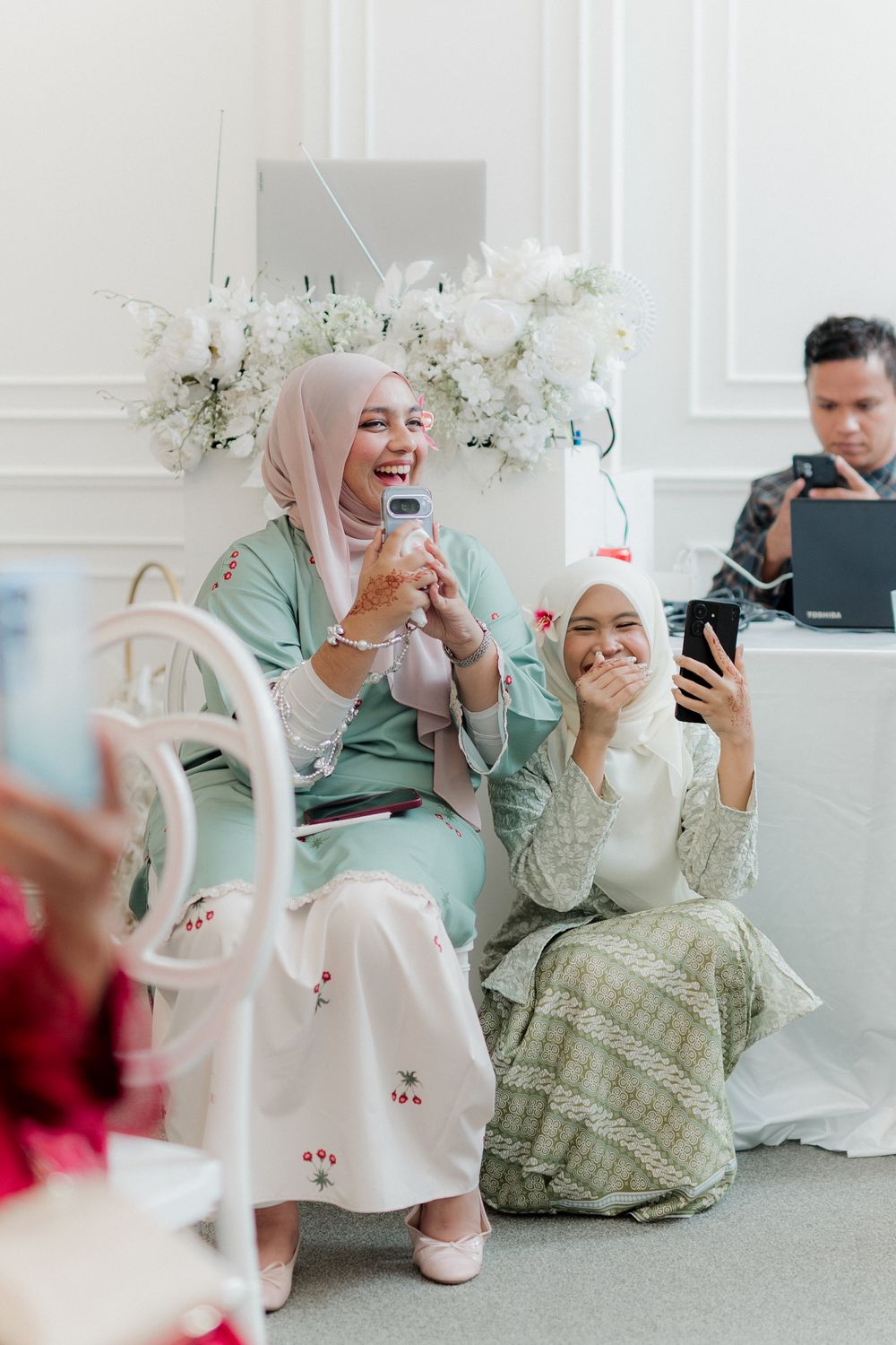 Two Muslim women in hijabs laugh joyfully at a elegant white floral event, holding phones while seated together.
