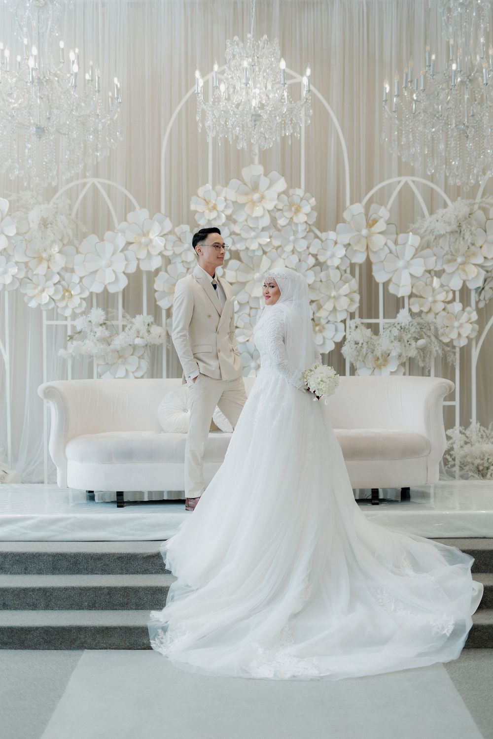 Muslim bride in white hijab gown and groom in beige suit pose before elegant floral wedding backdrop.