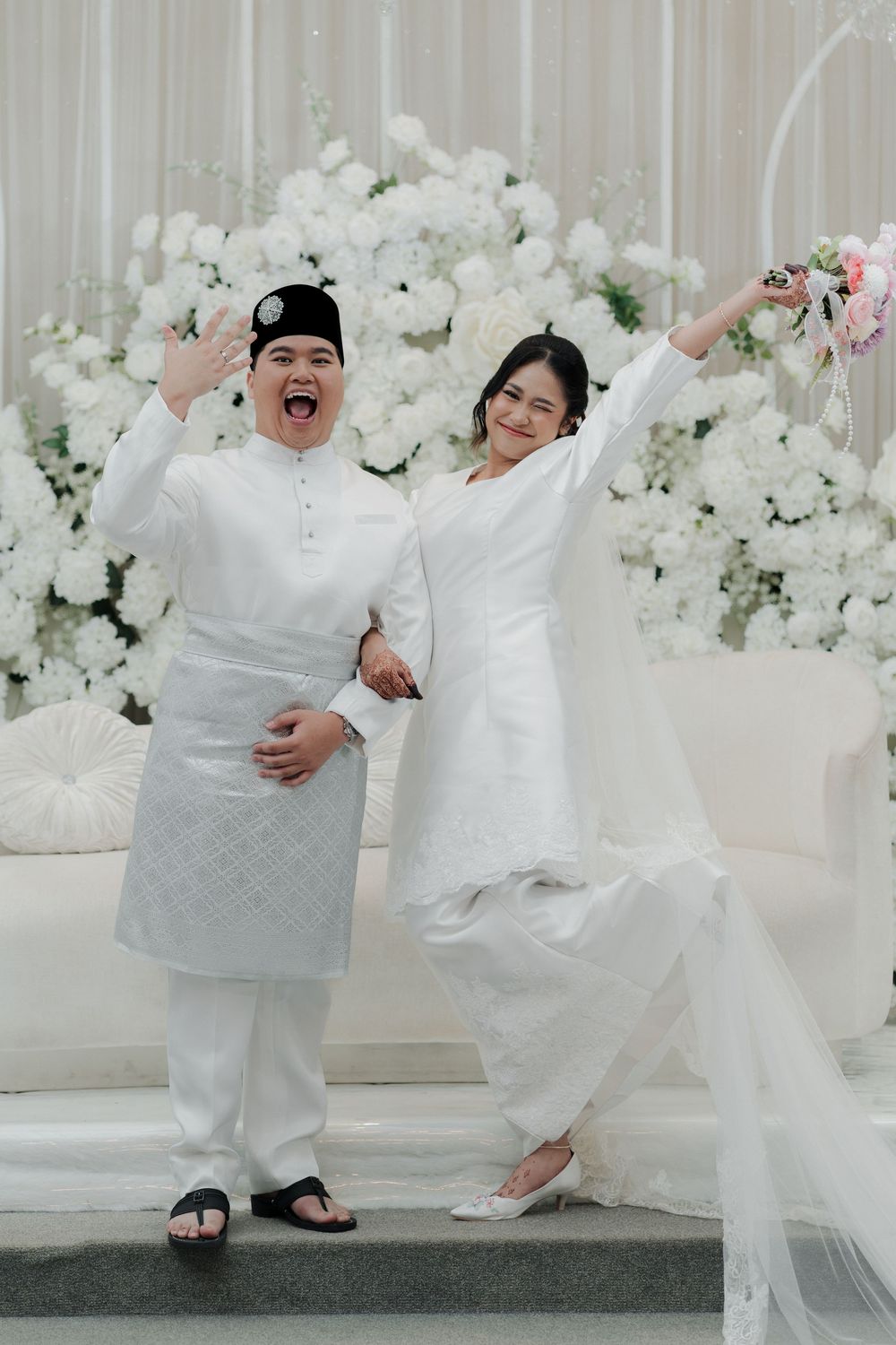 Joyful couple in white traditional Malay attire celebrating their wedding day in front of white floral backdrop.