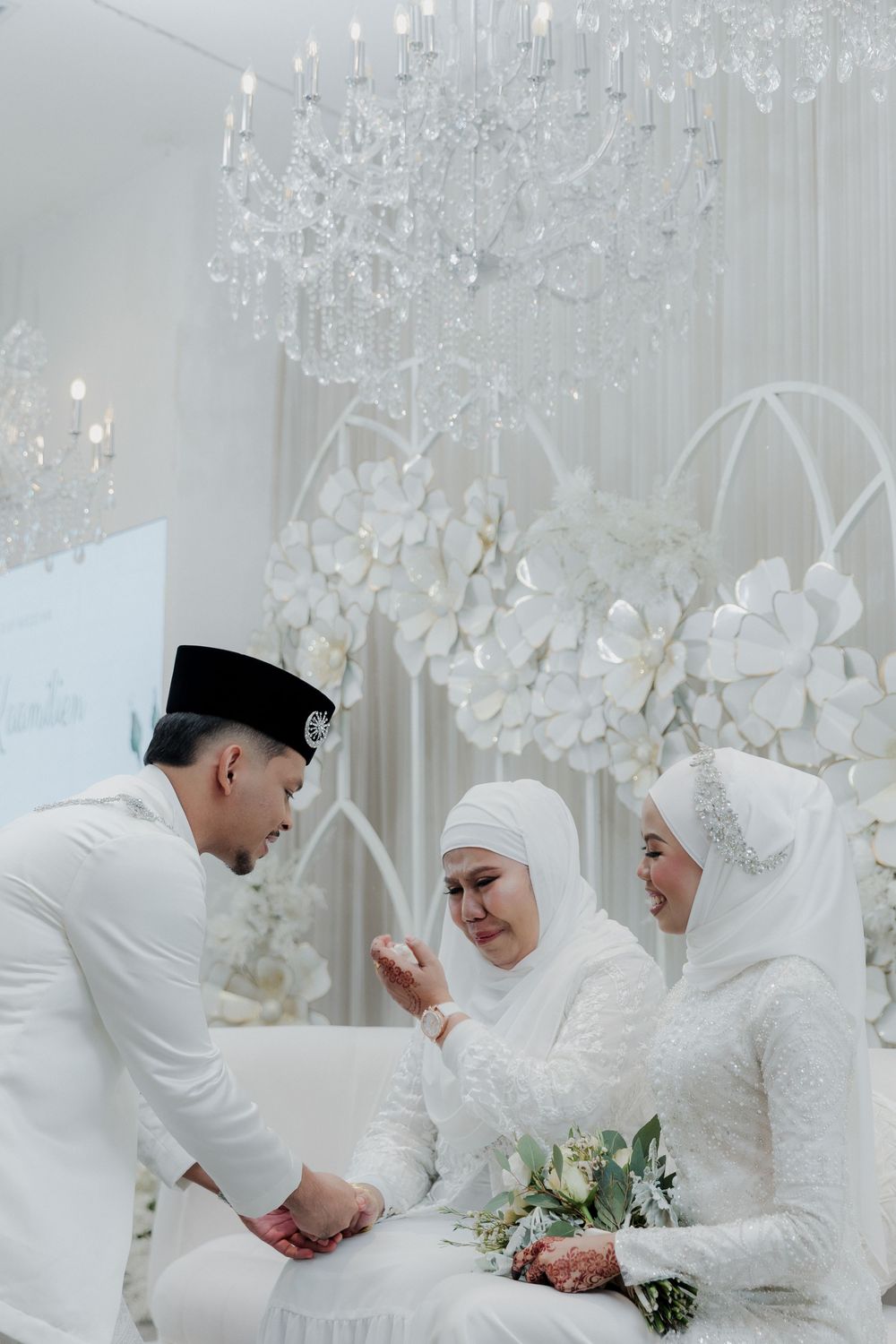 Muslim wedding ceremony with bride, groom, and guest in white attire under crystal chandelier with floral backdrop.