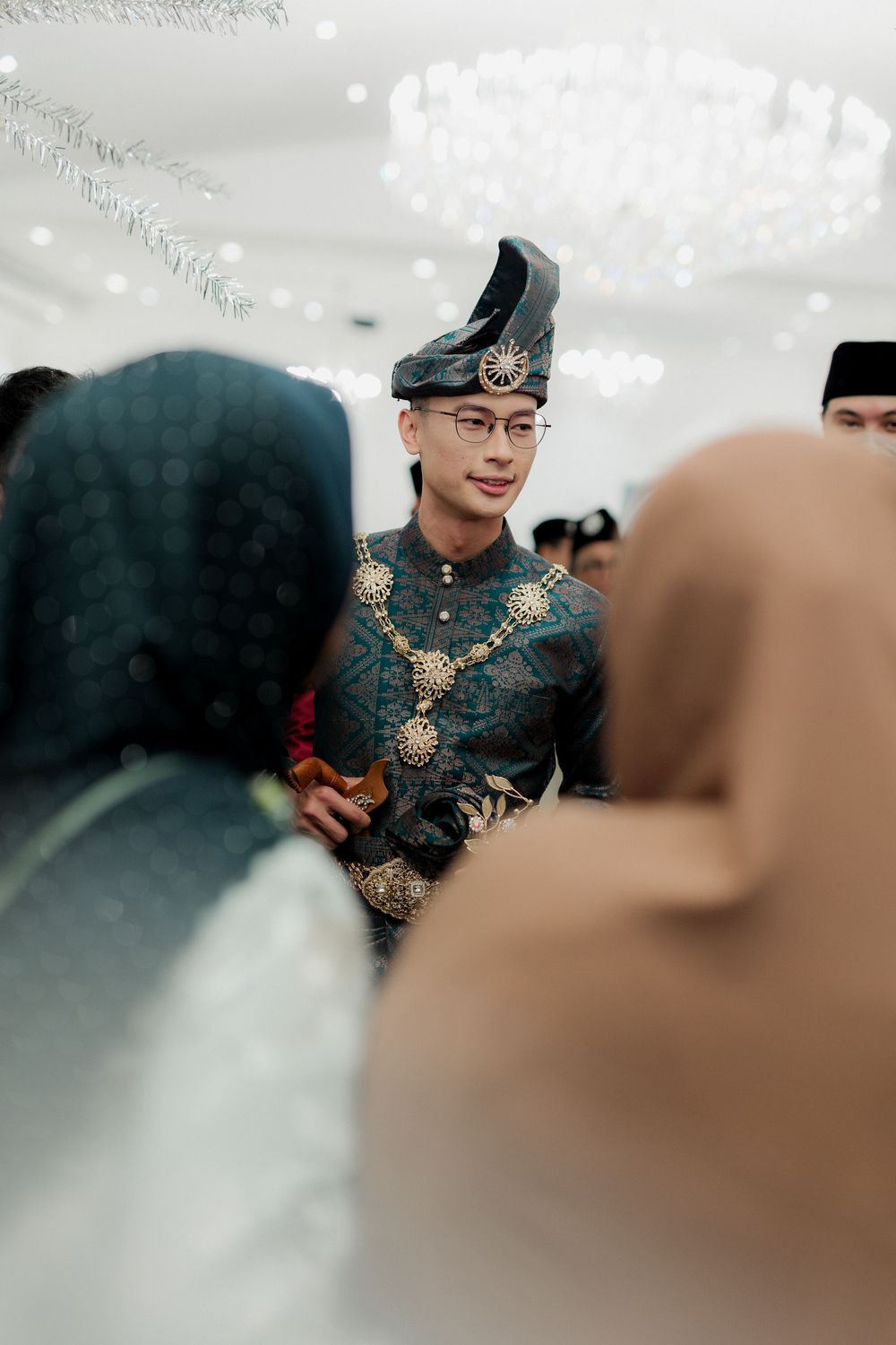 Groom in traditional Malay attire with gold accessories and songkok hat greeting guests at a wedding ceremony.